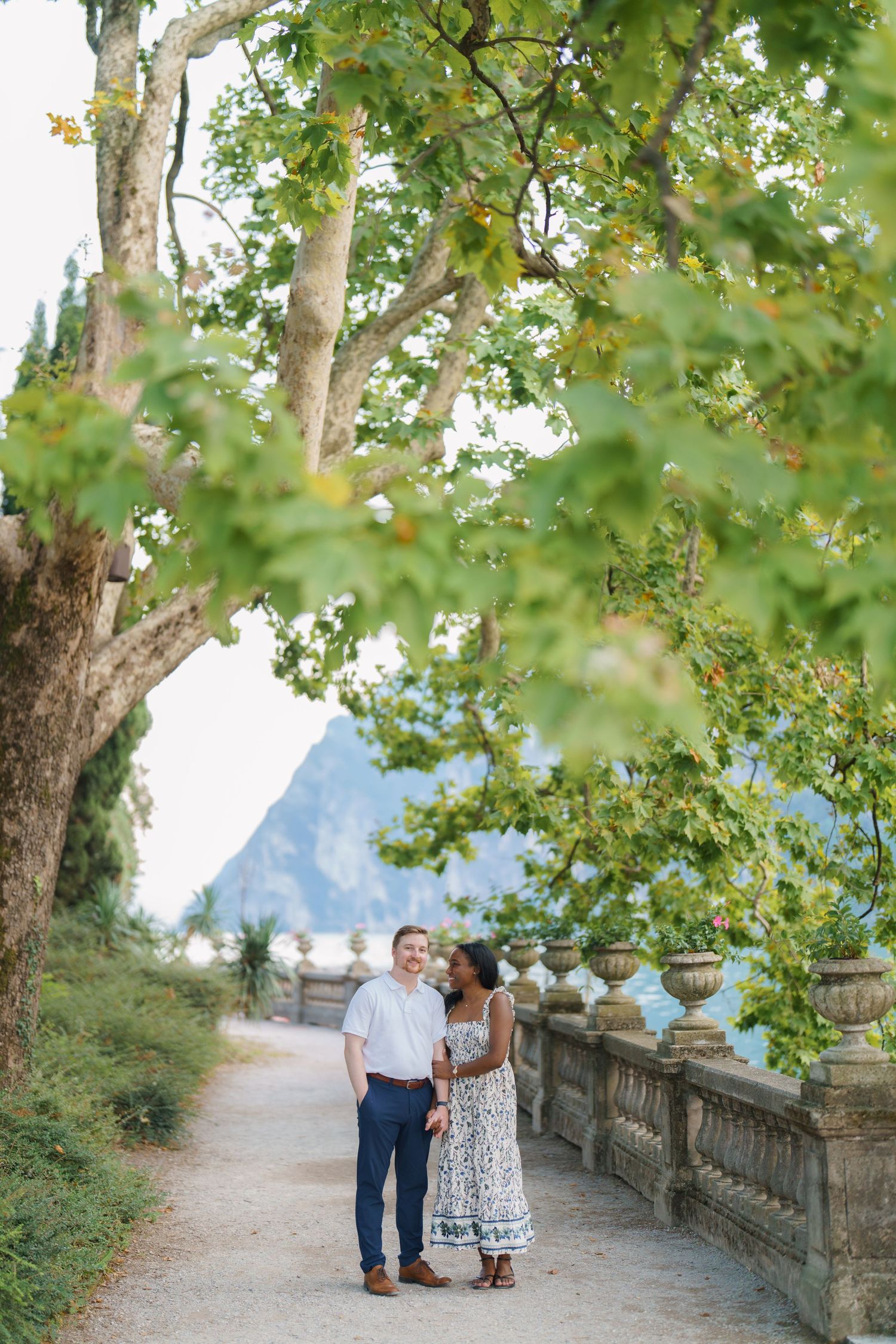 Couple walking hand in hand along a tree-lined stone path with mountain and lake scenery.