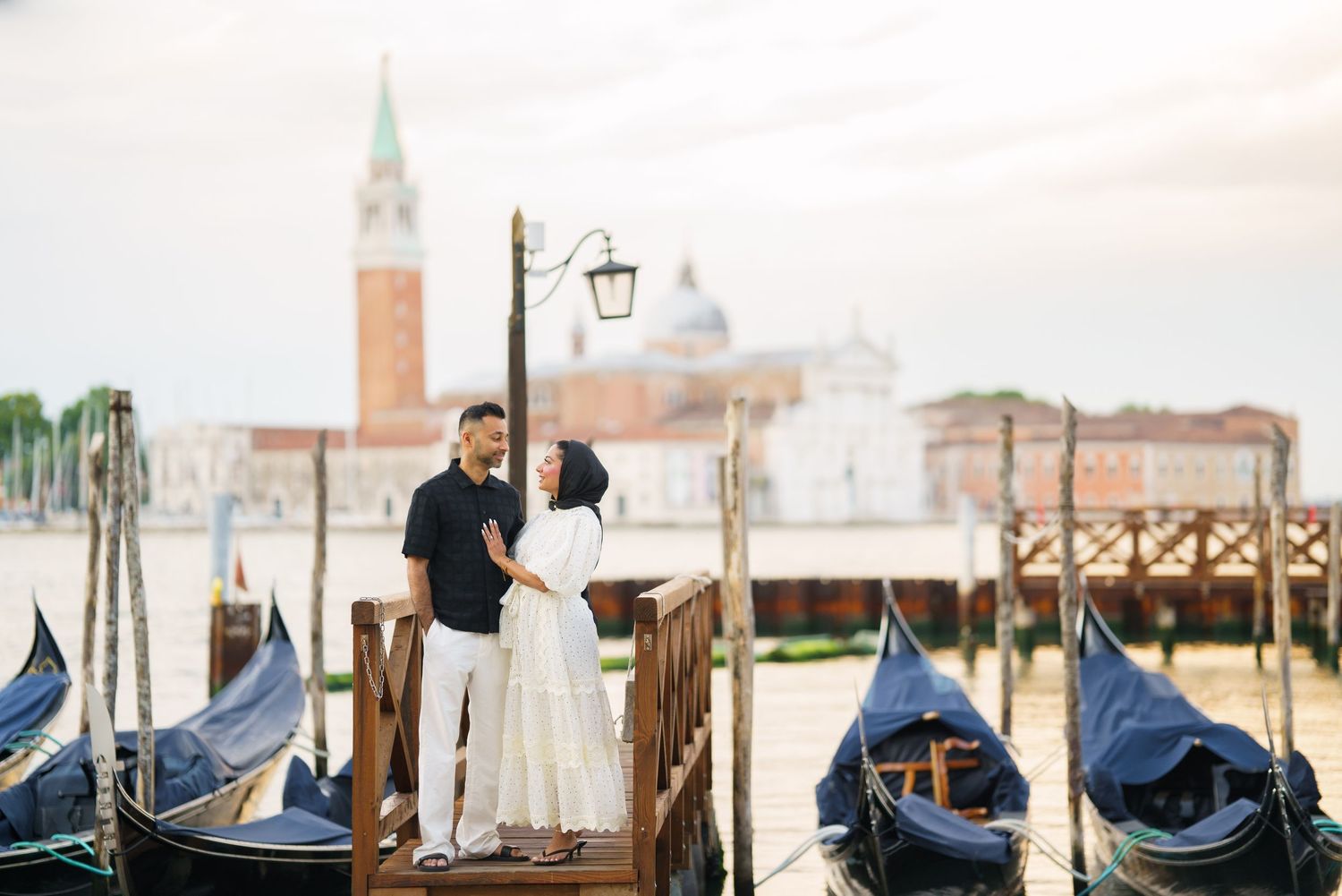 Couple embracing on a wooden dock in Venice, Italy, with gondolas and San Giorgio Maggiore island in the background.