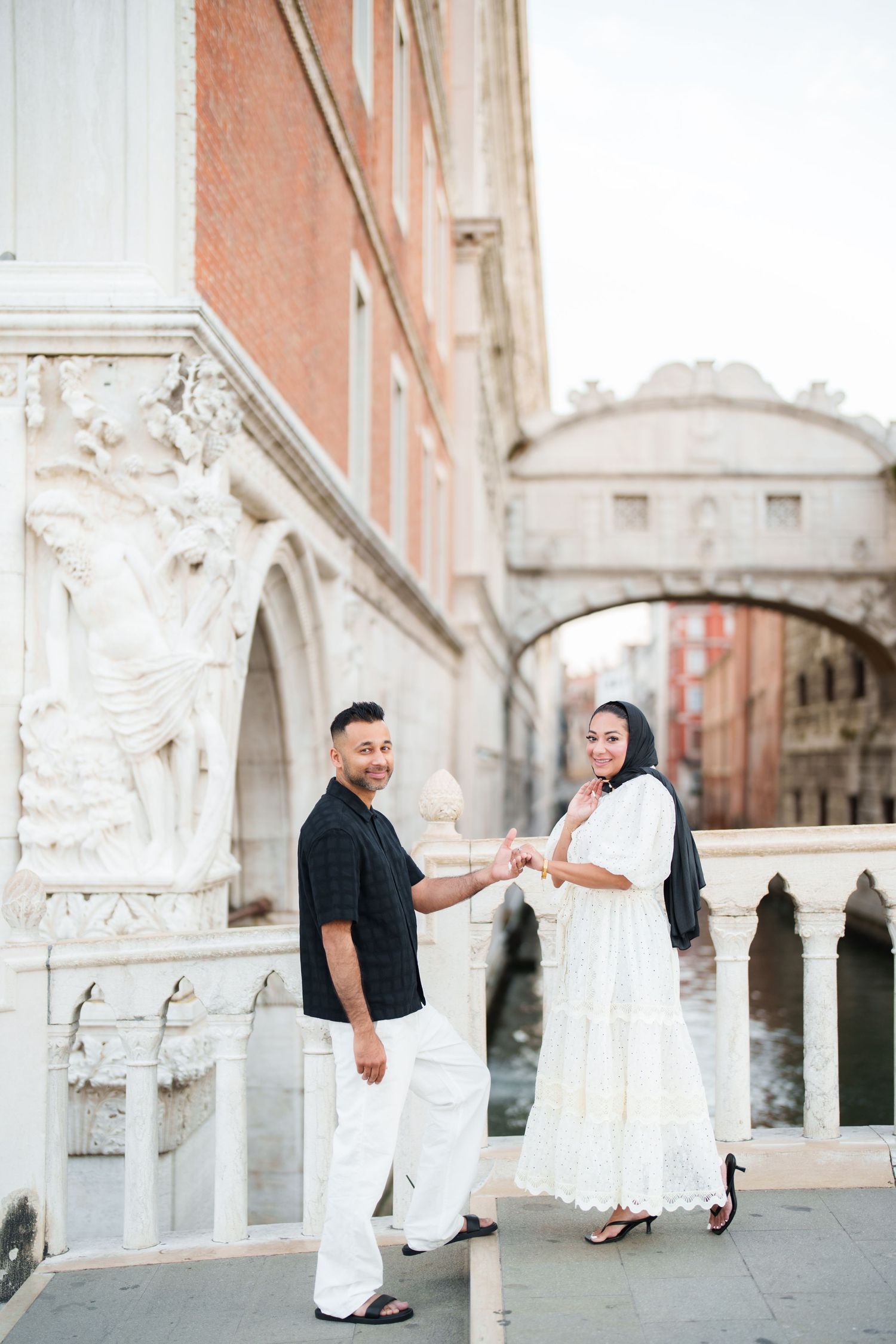 Couple holding hands on a bridge in Venice, Italy, with the Bridge of Sighs in the background.