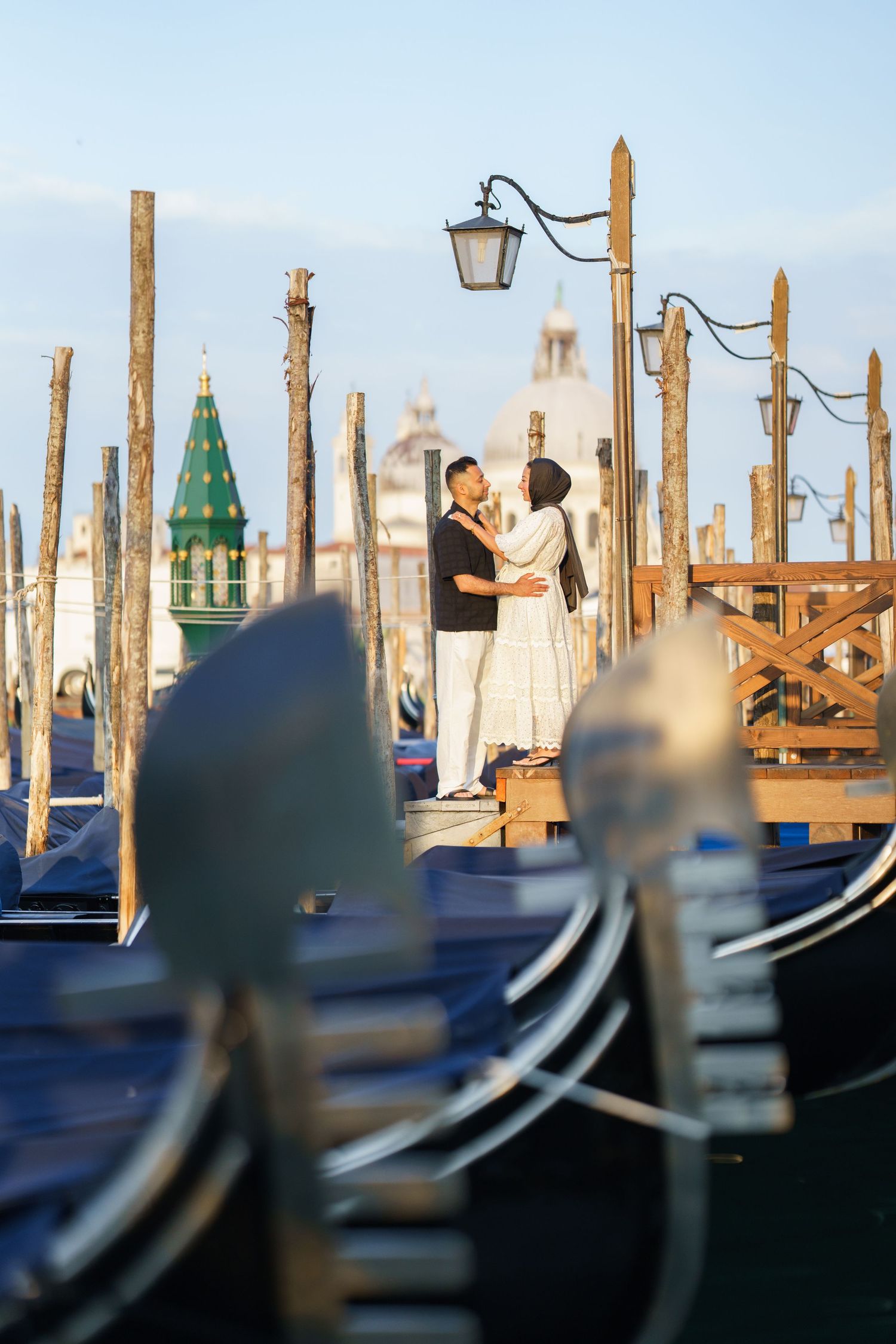Couple embracing on Venice gondola dock with wooden poles and historic architecture in soft golden light.