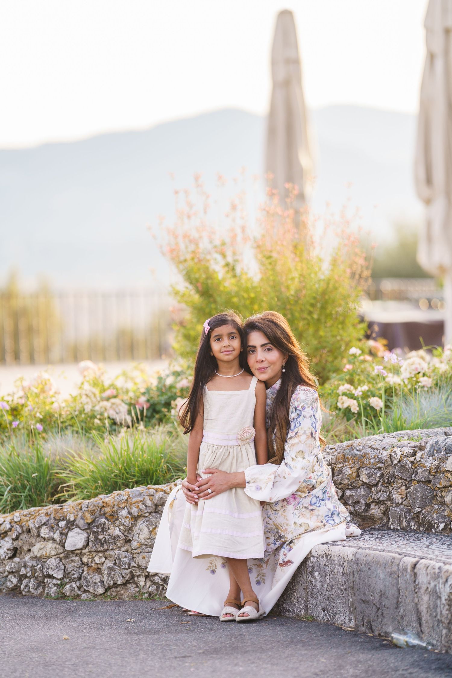 A woman and young girl in white dresses pose together on a stone bench with mountains and flowers behind them.