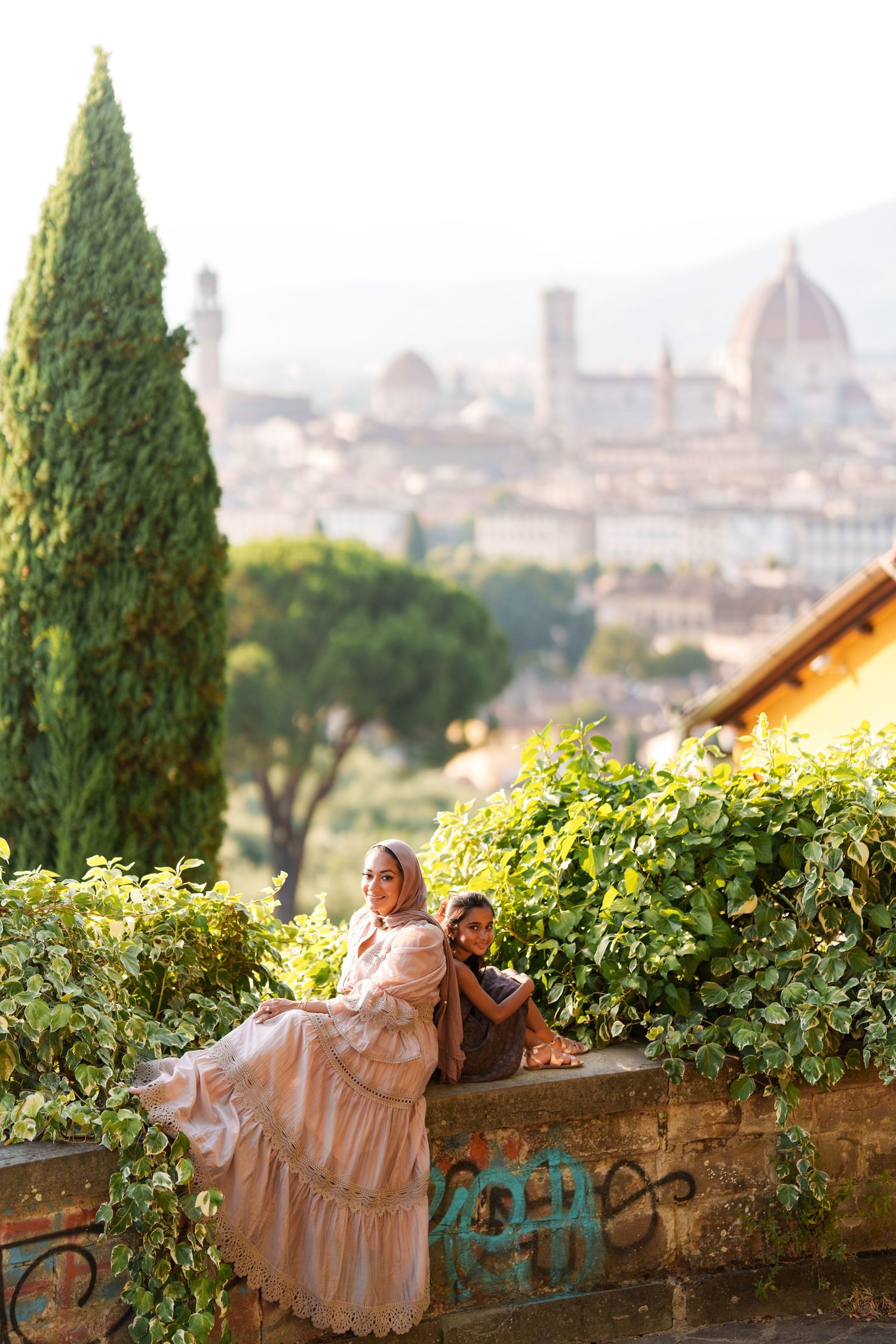 Woman in pink dress and child sit on stone wall overlooking Florence skyline with Duomo cathedral backdrop.