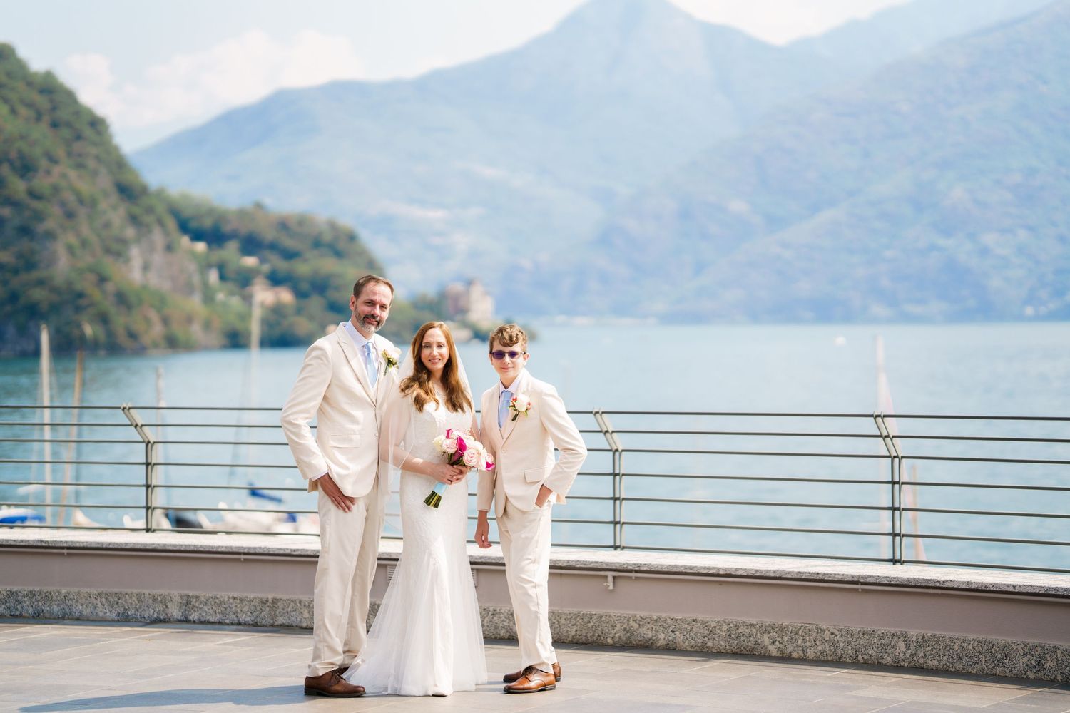 Bride and groom with officiant posing on lakeside terrace with mountain backdrop at outdoor wedding ceremony.