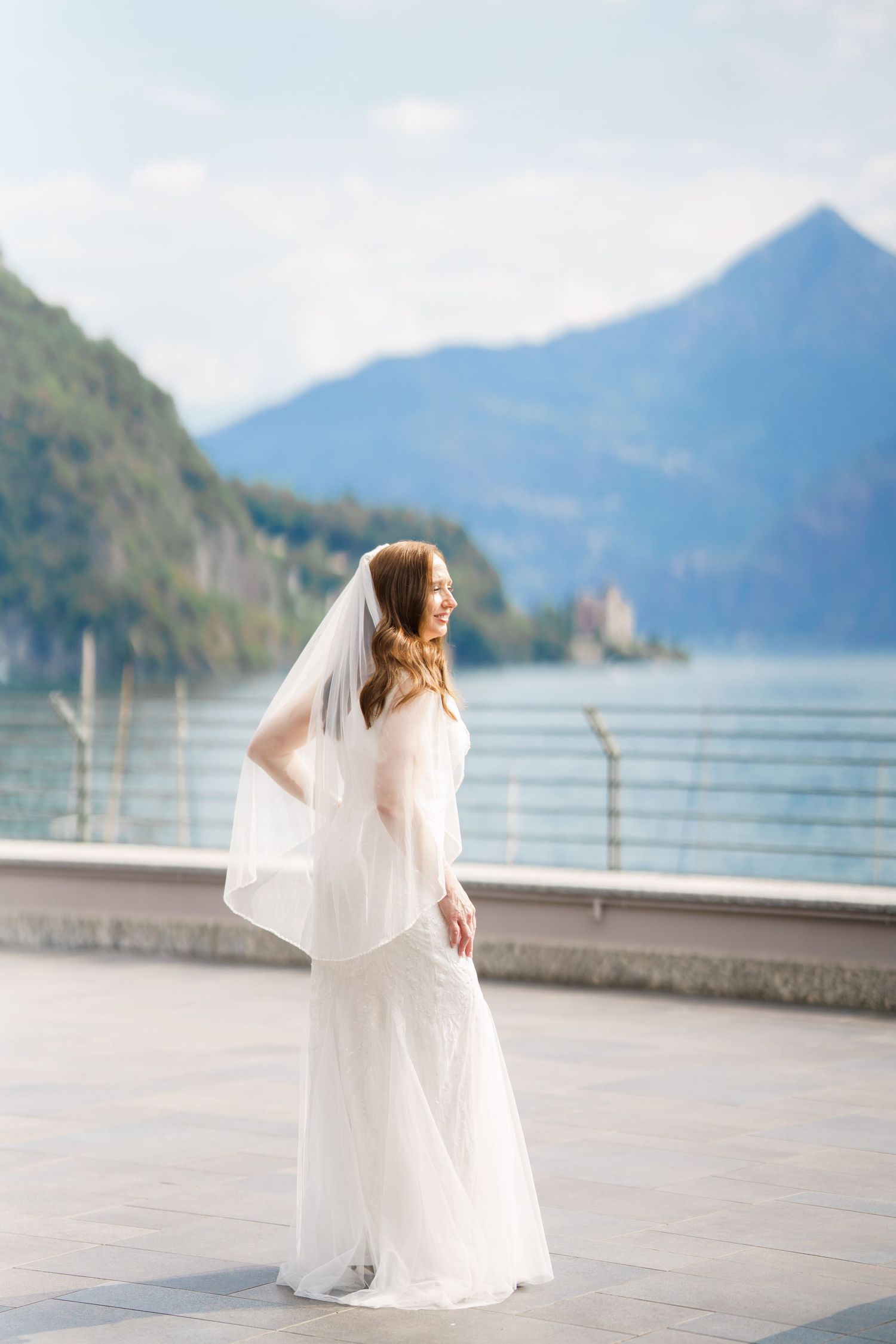 Smiling bride in white gown and veil stands on terrace with mountain lake backdrop on her wedding day.