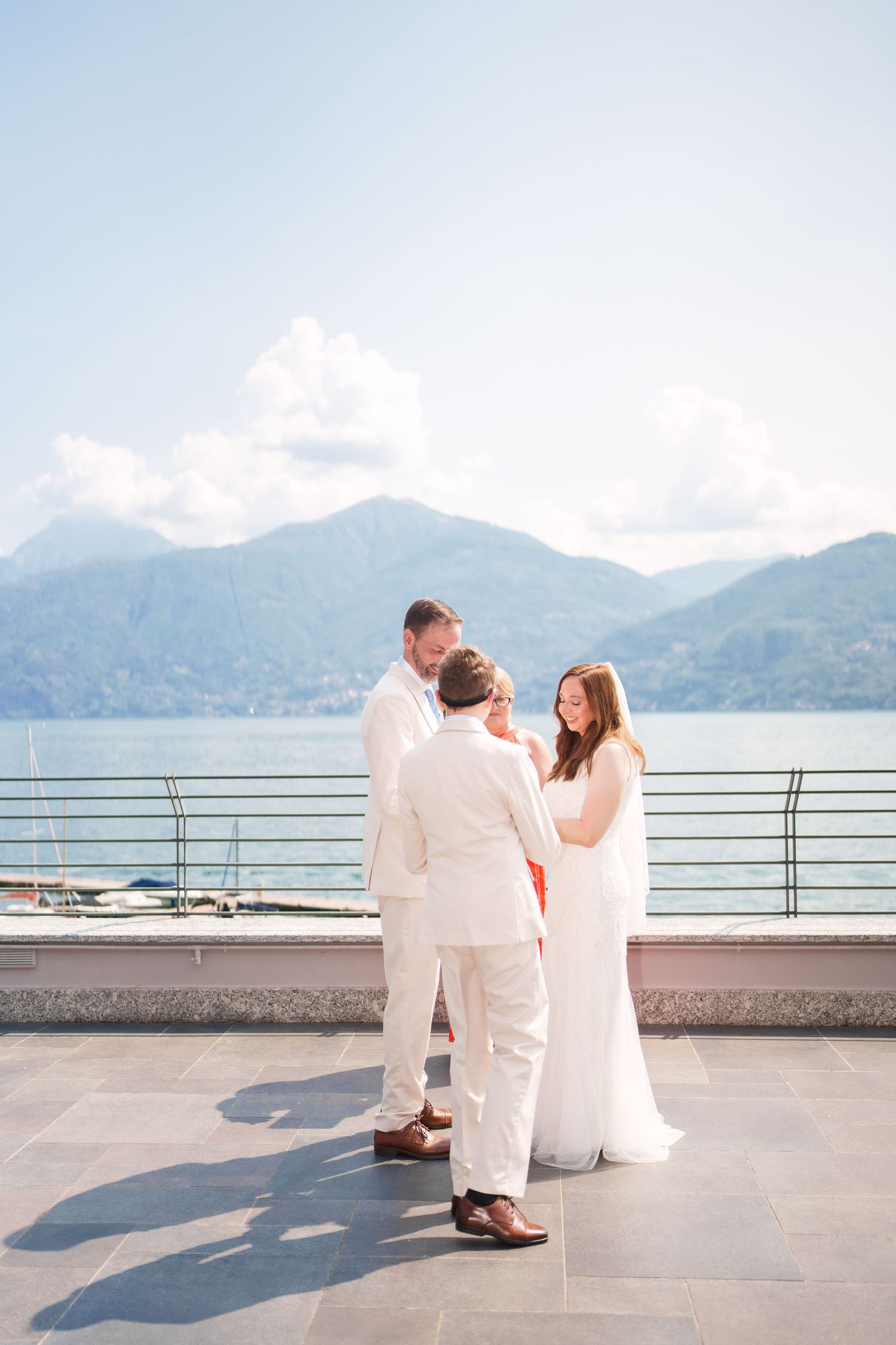 Couple exchanges vows with officiant on a sunlit terrace overlooking Lake Como with mountains in the background.