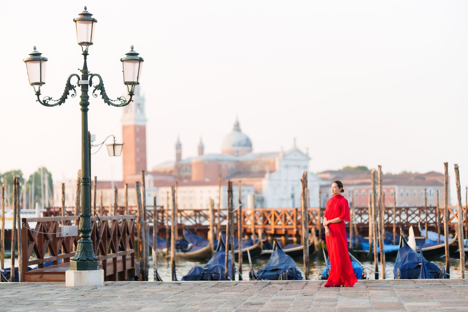 Woman in red dress standing near gondolas and lamp post in Venice, Italy with San Giorgio Maggiore in background.