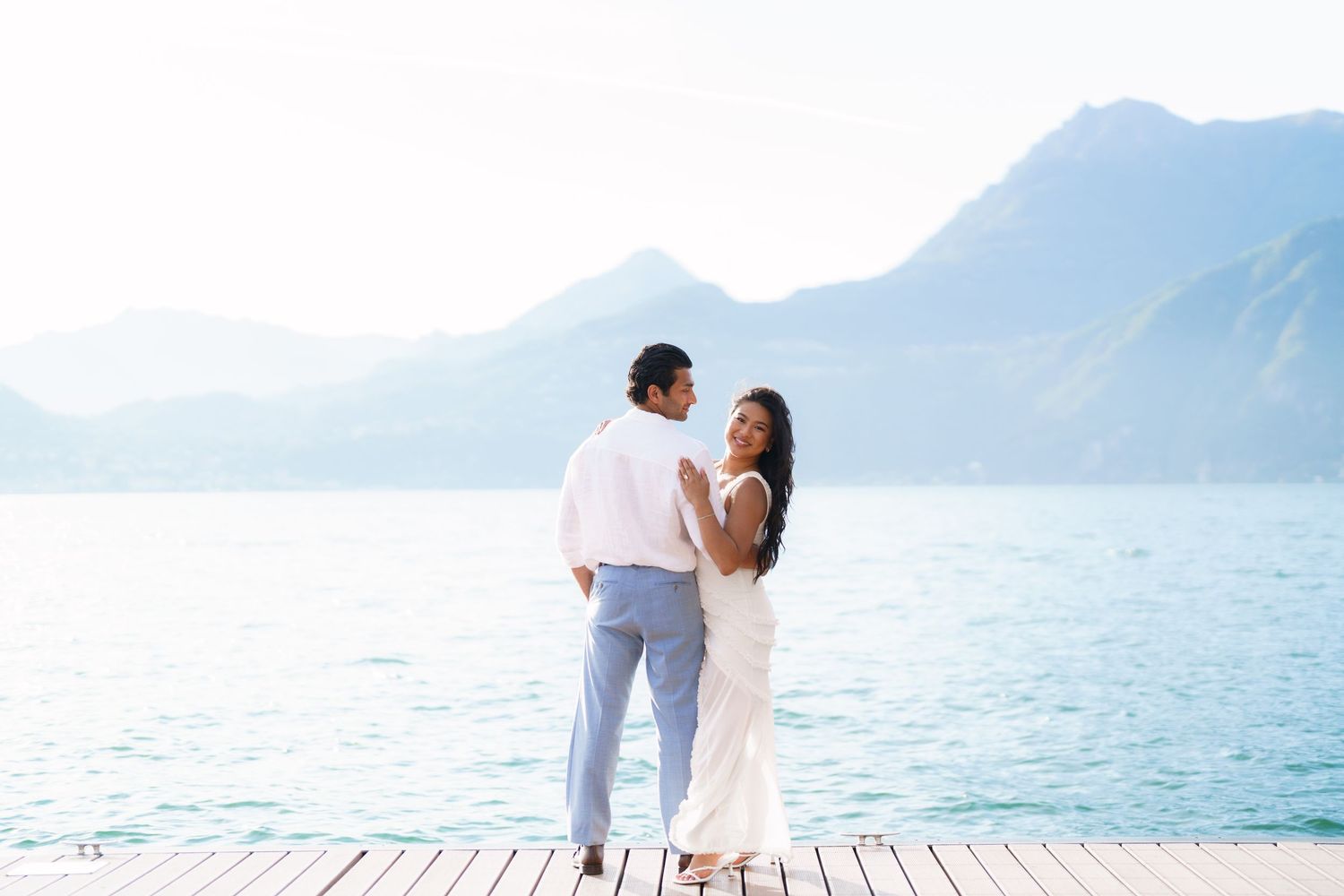 Couple in white attire standing on a dock embracing with serene blue lake and misty mountains in background.
