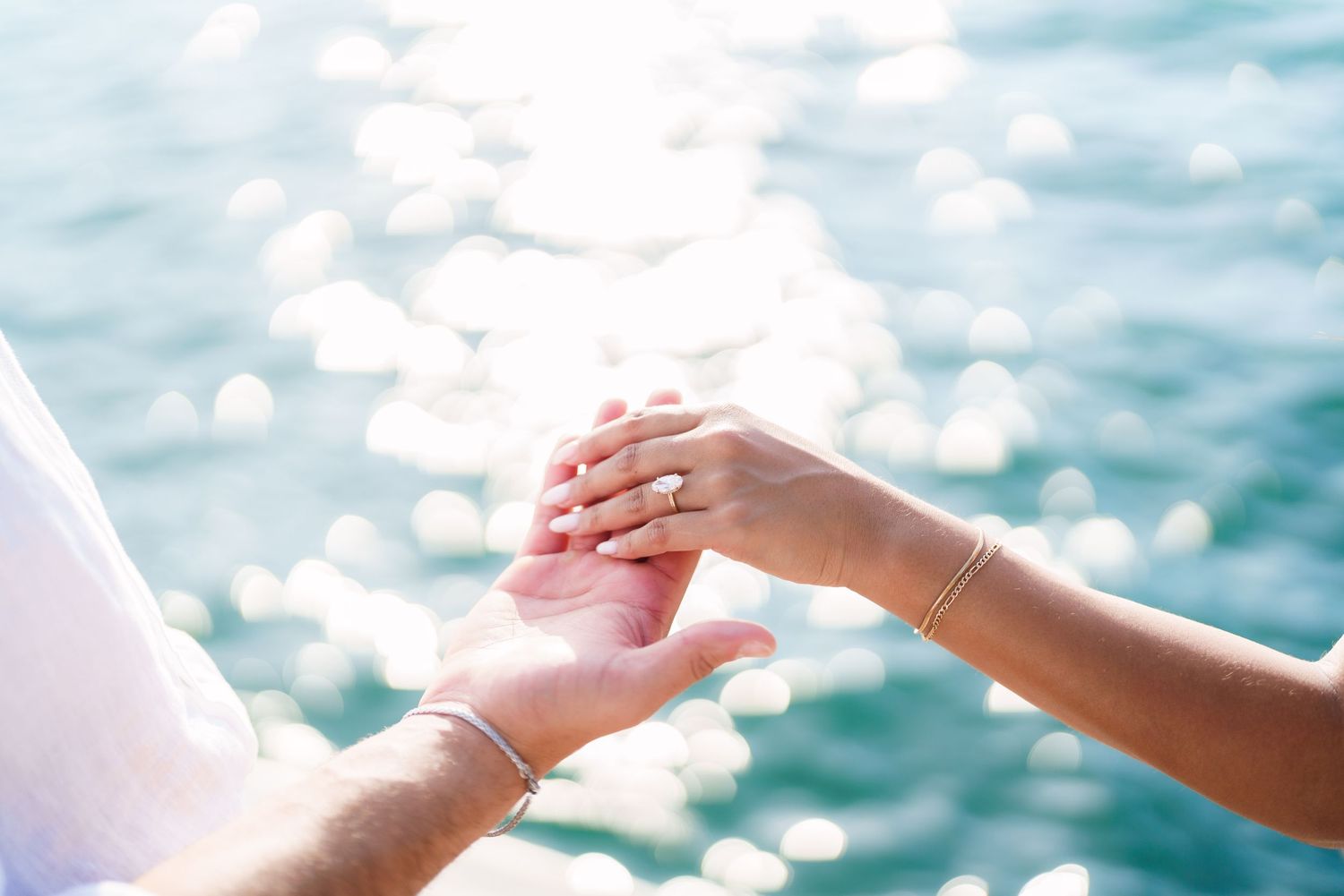 Two people holding hands with sparkling turquoise ocean water in the background on a sunny day.
