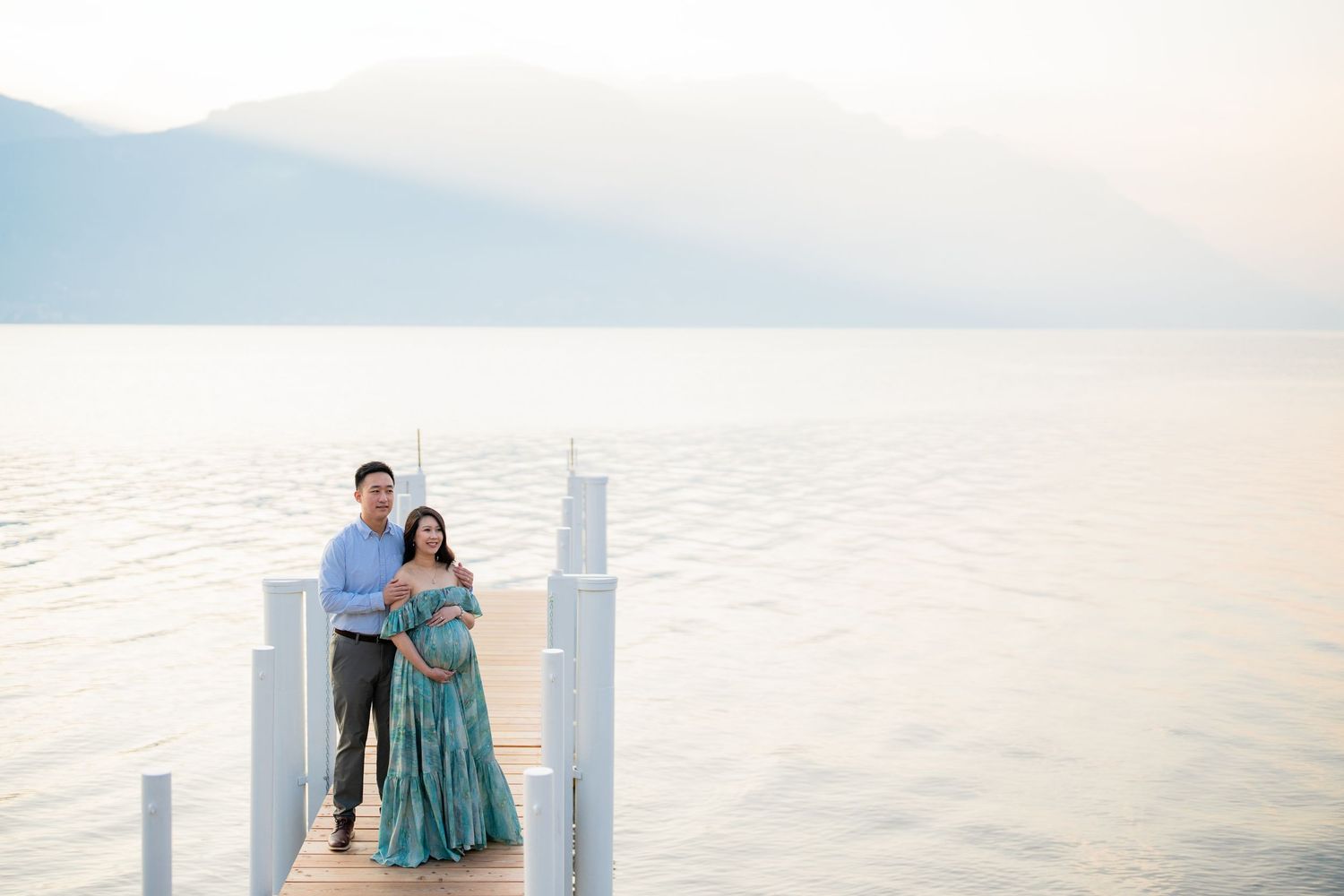 Couple poses on a lakeside dock at sunset, woman in teal maternity gown, mountains in background.