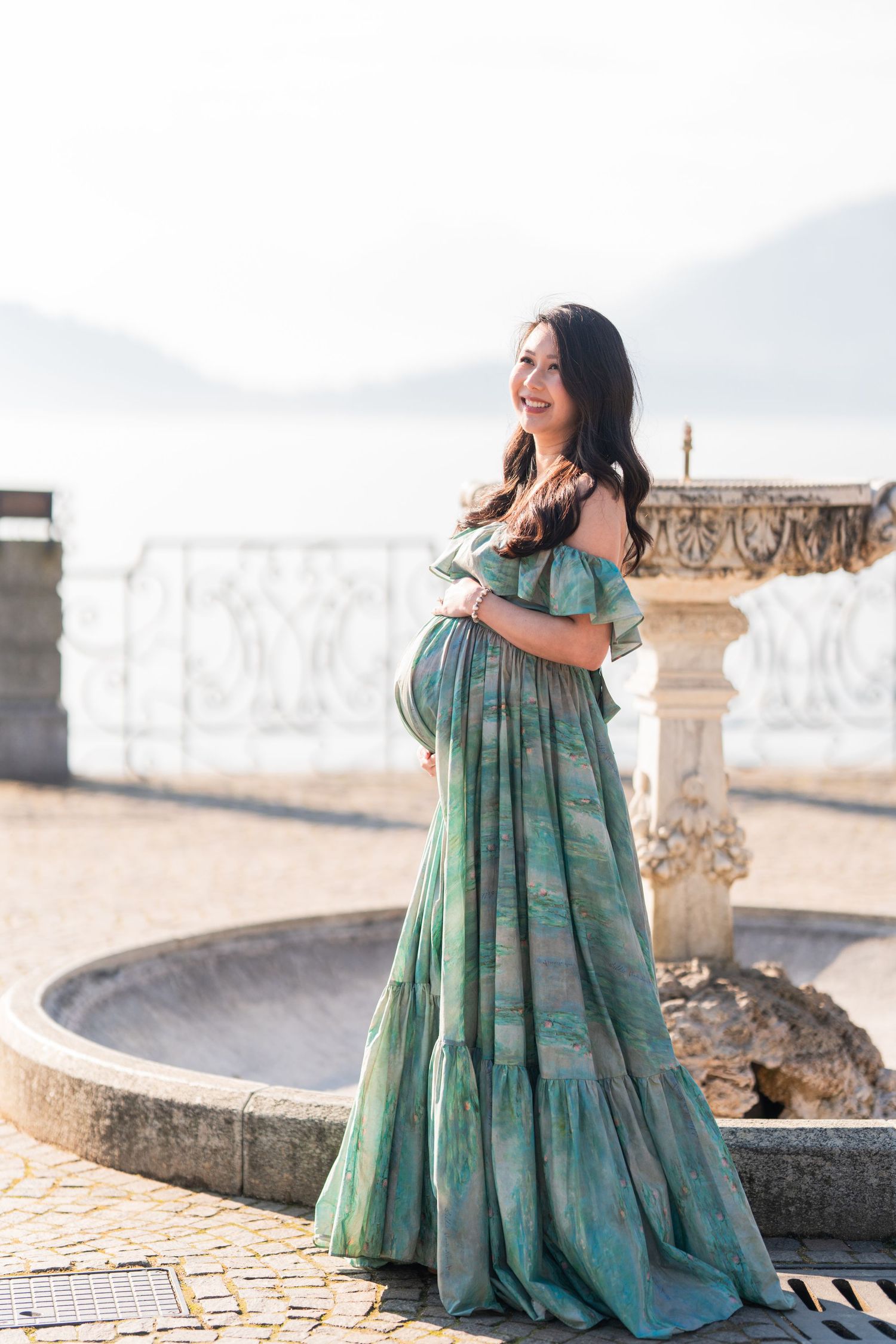Smiling pregnant woman in green off-shoulder floral gown standing by stone fountain with mountain lake backdrop.