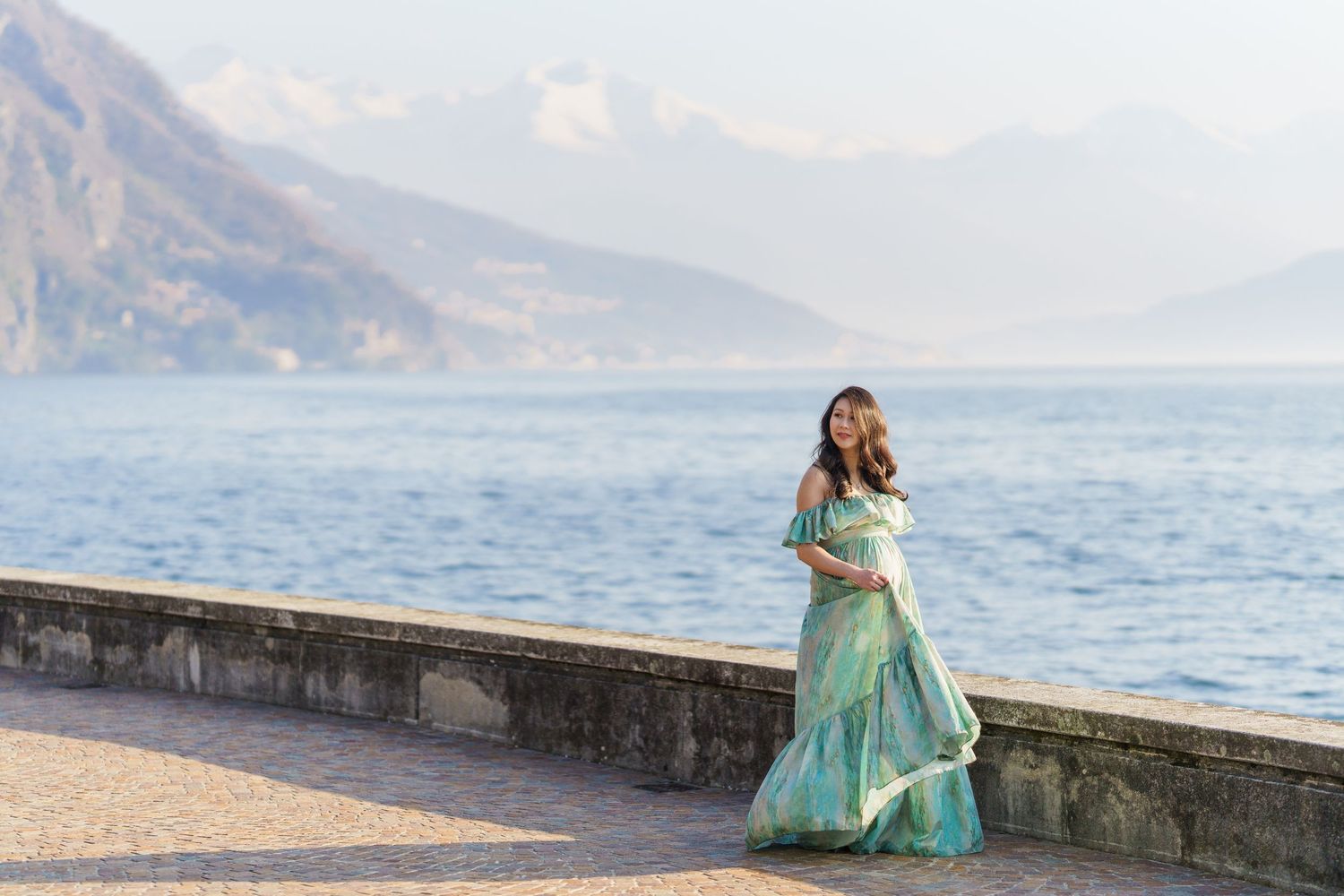 Pregnant woman in flowing teal gown poses by Lake Como waterfront with misty mountains backdrop.