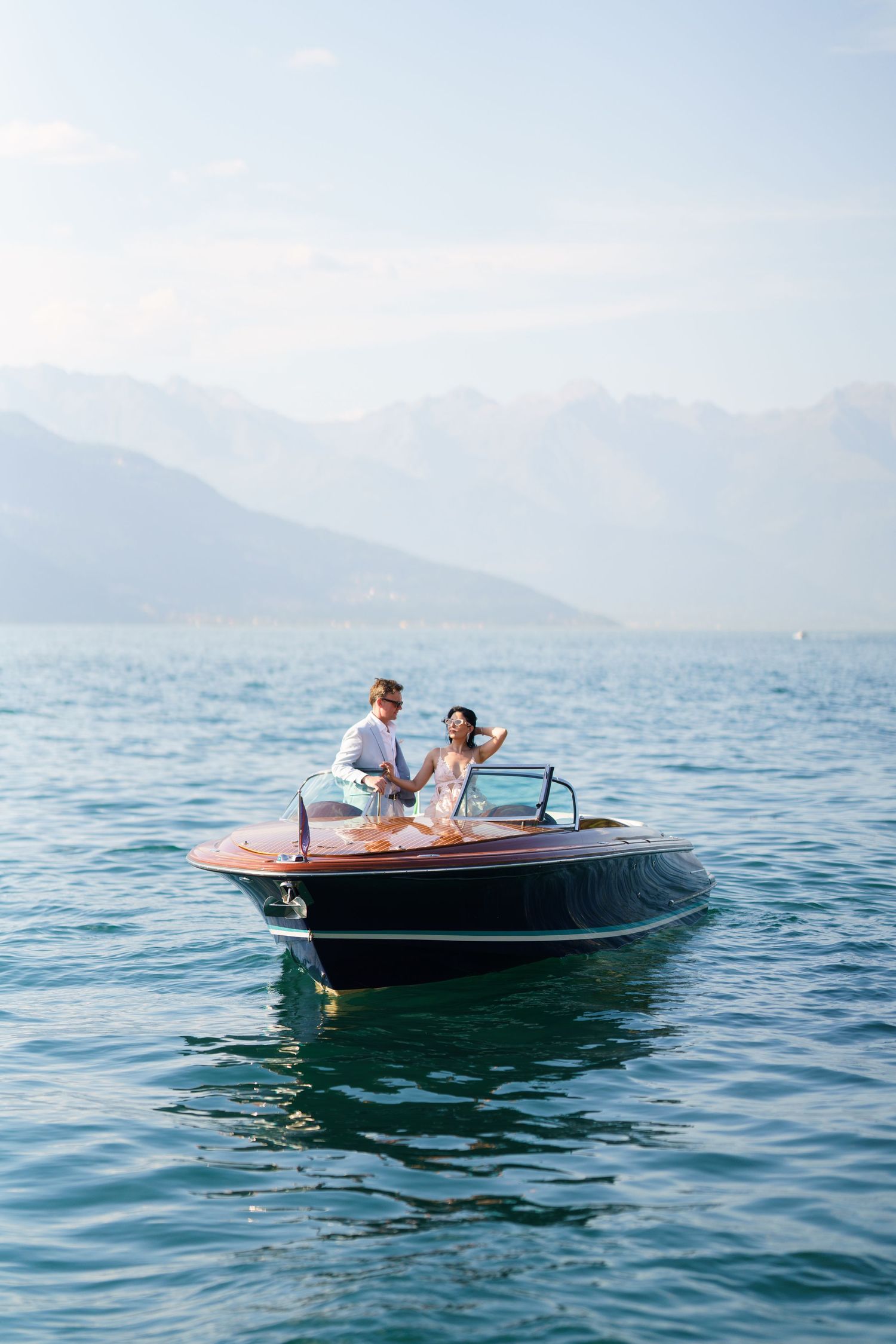Couple in wedding attire riding a classic wooden speedboat on a calm blue lake with misty mountains in the background.
