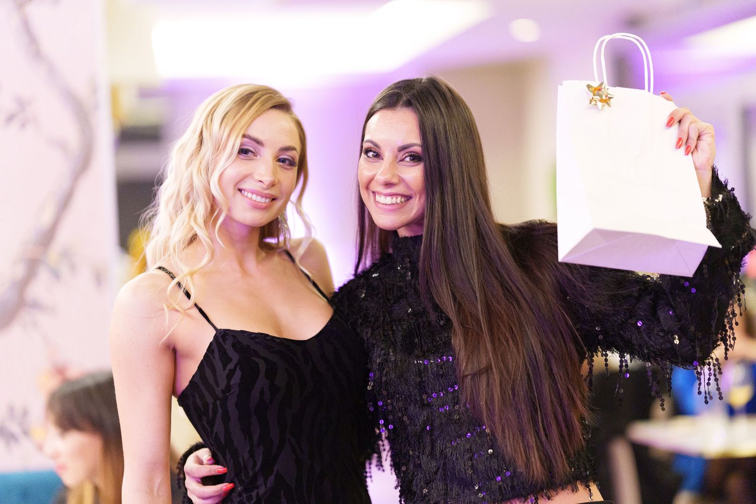 Two women in elegant black outfits pose together, one holding a white gift bag at an upscale event.