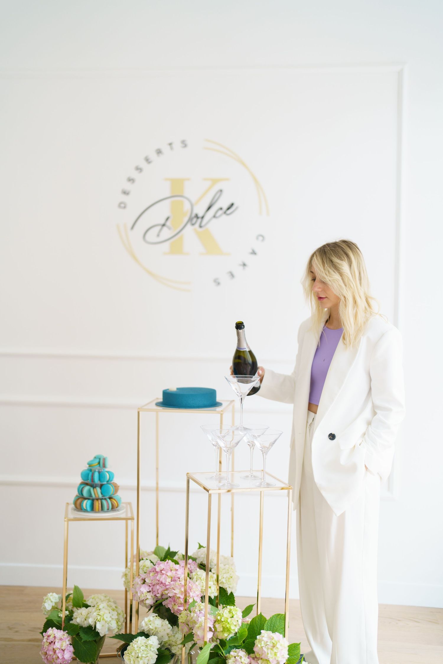 Woman in white suit pouring champagne at Dolce K Desserts elegant display with macarons and hydrangeas.