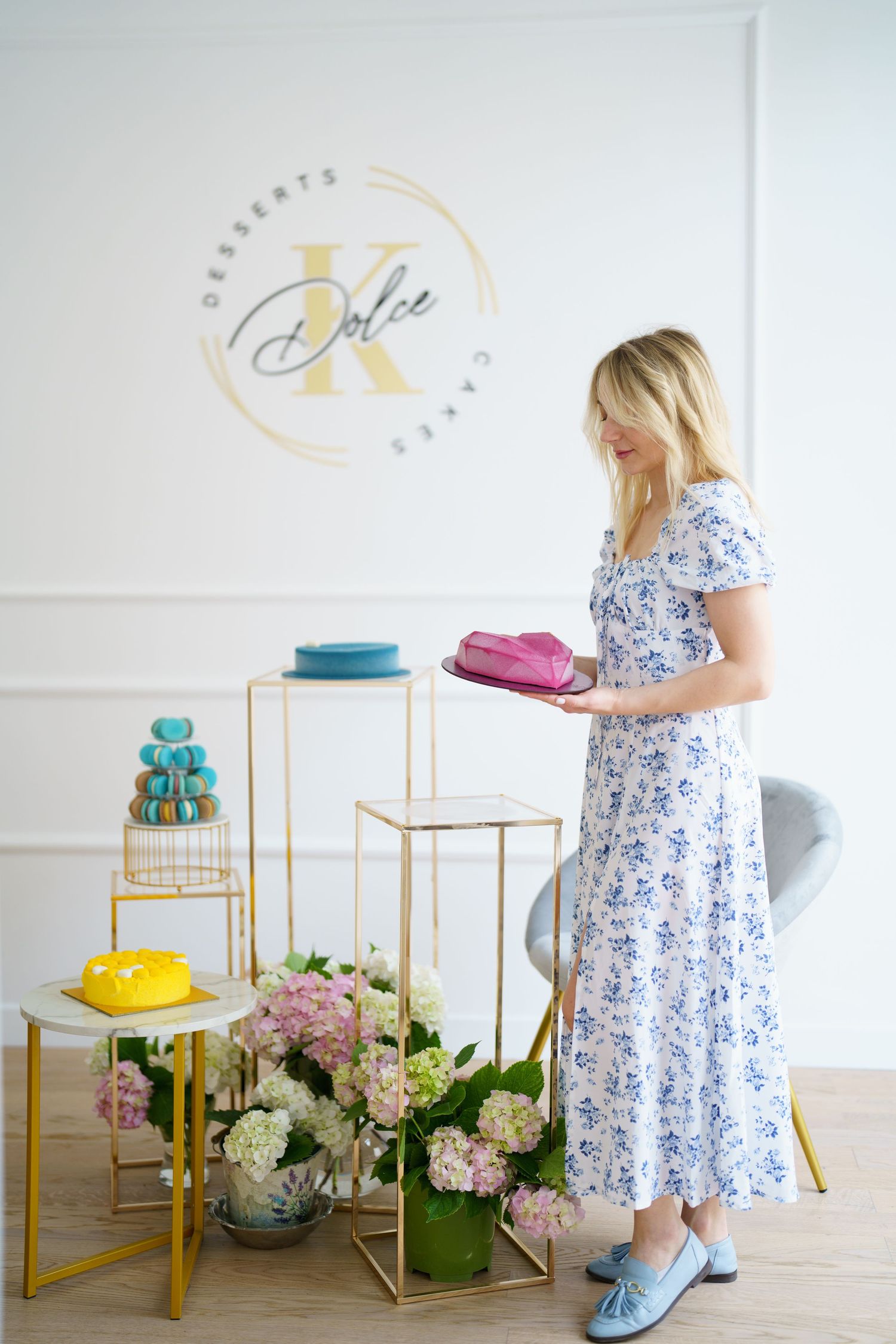 Woman in floral dress holding pink cake at K Dolce Desserts & Cakes shop with colorful macarons and hydrangeas.