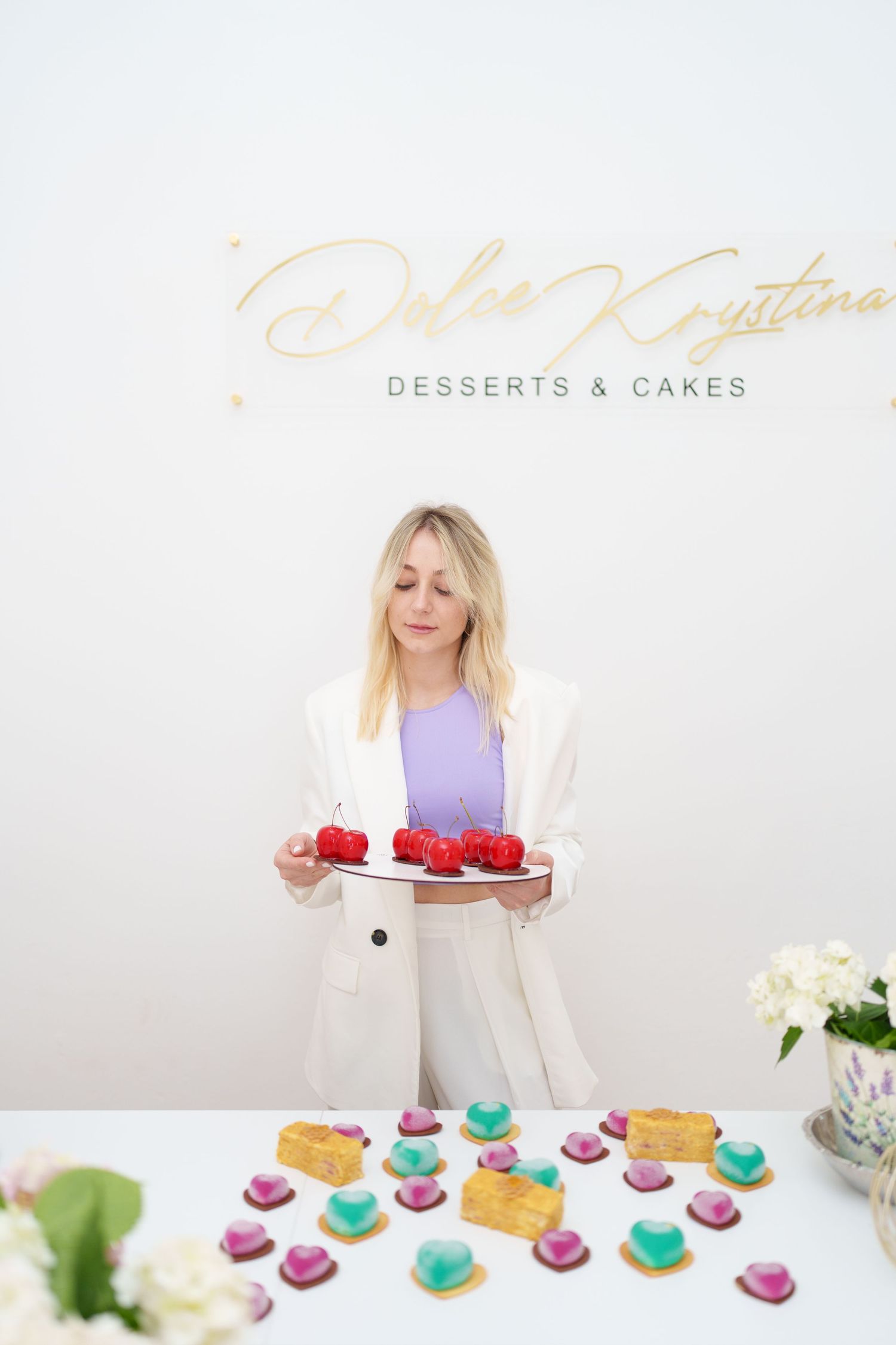 Woman in white blazer holding red cake at Dolce Krystina Desserts & Cakes shop with colorful treats on table.