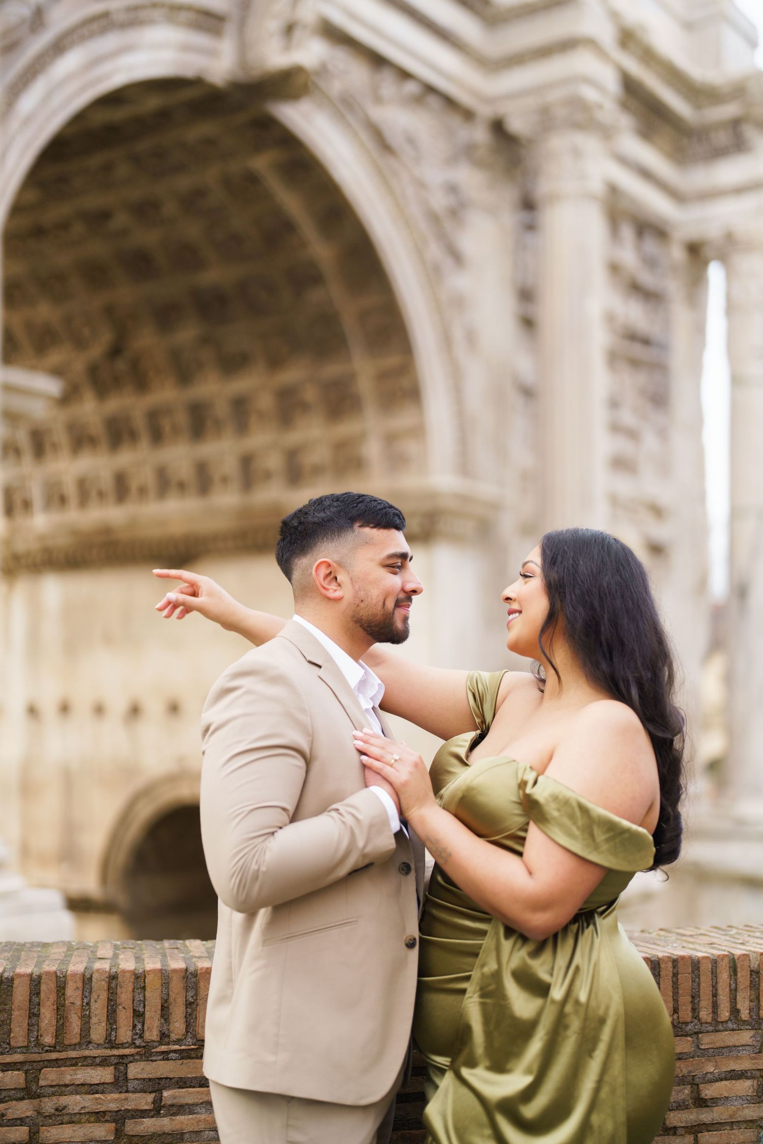 Couple in elegant attire embracing and smiling before a grand stone arch monument.