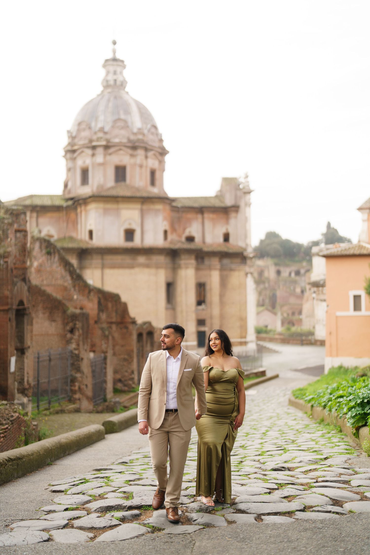 Couple in elegant attire walking on cobblestone path with Roman church dome backdrop in Rome, Italy.