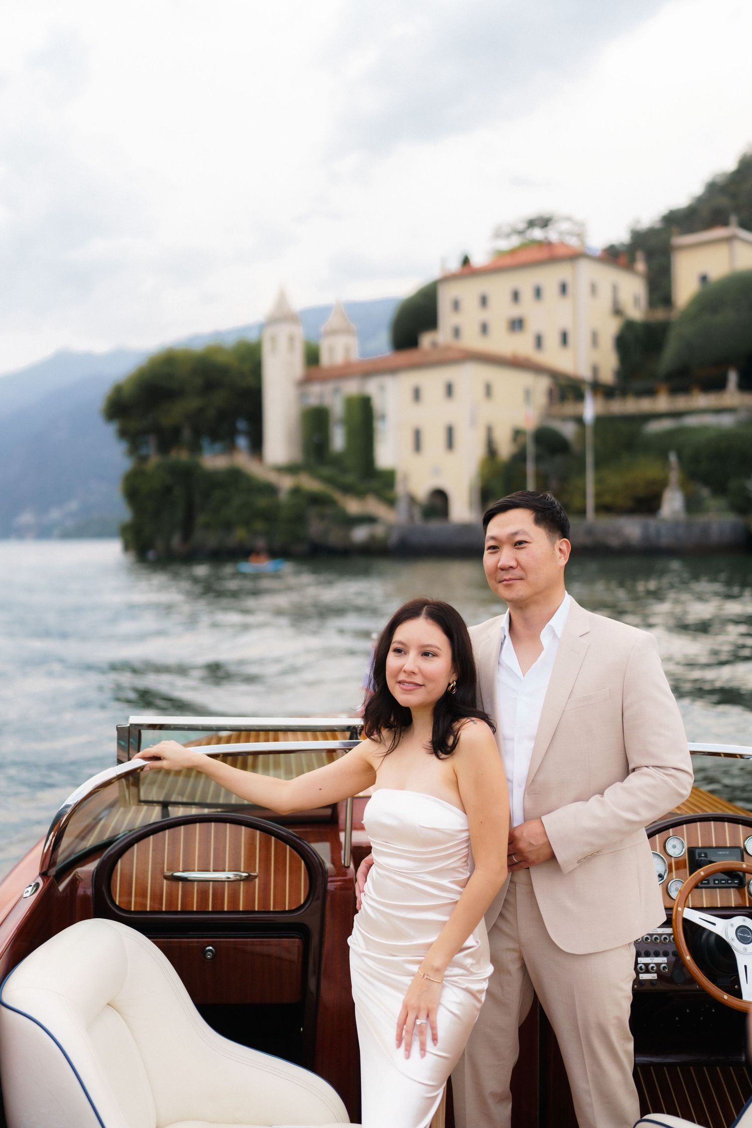 Couple in elegant white attire pose on wooden boat at Lake Como with Villa del Balbianello in background.