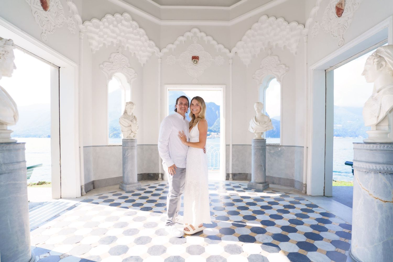 Romantic couple embraces in an elegant white marble pavilion with checkerboard floor overlooking the ocean.