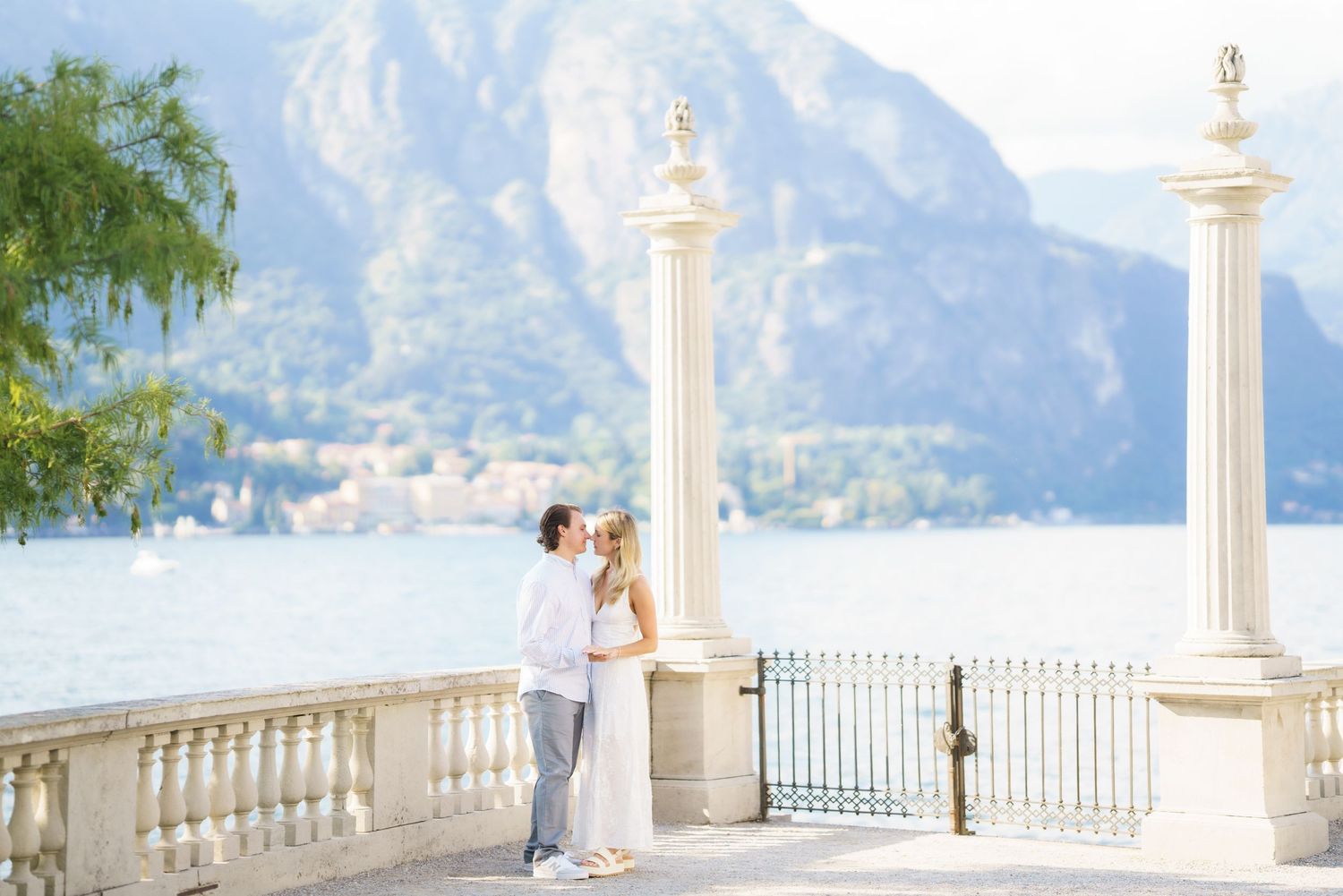Romantic couple embracing on elegant lakeside terrace with white pillars and mountain backdrop in Lake Como, Italy.