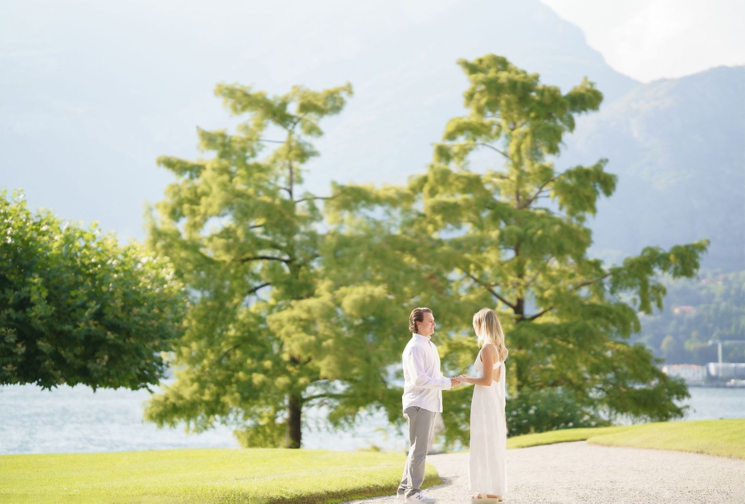 A couple in white outfits walks together along a scenic lakeside path with mountains and trees in the background.
