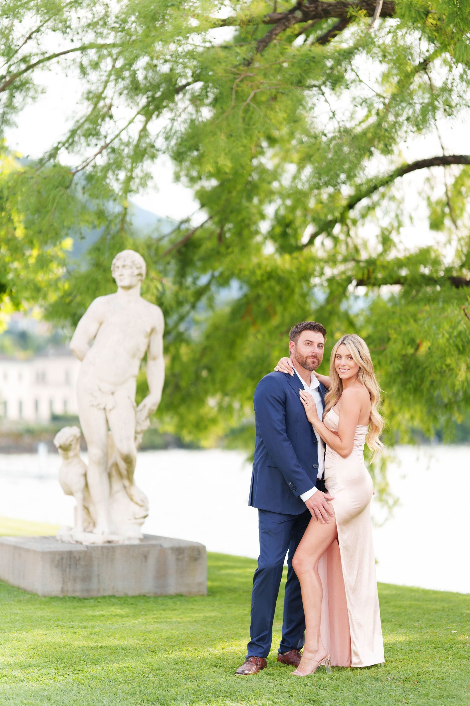 A romantic couple in formal attire poses for photos near a stone statue in a lush garden setting.