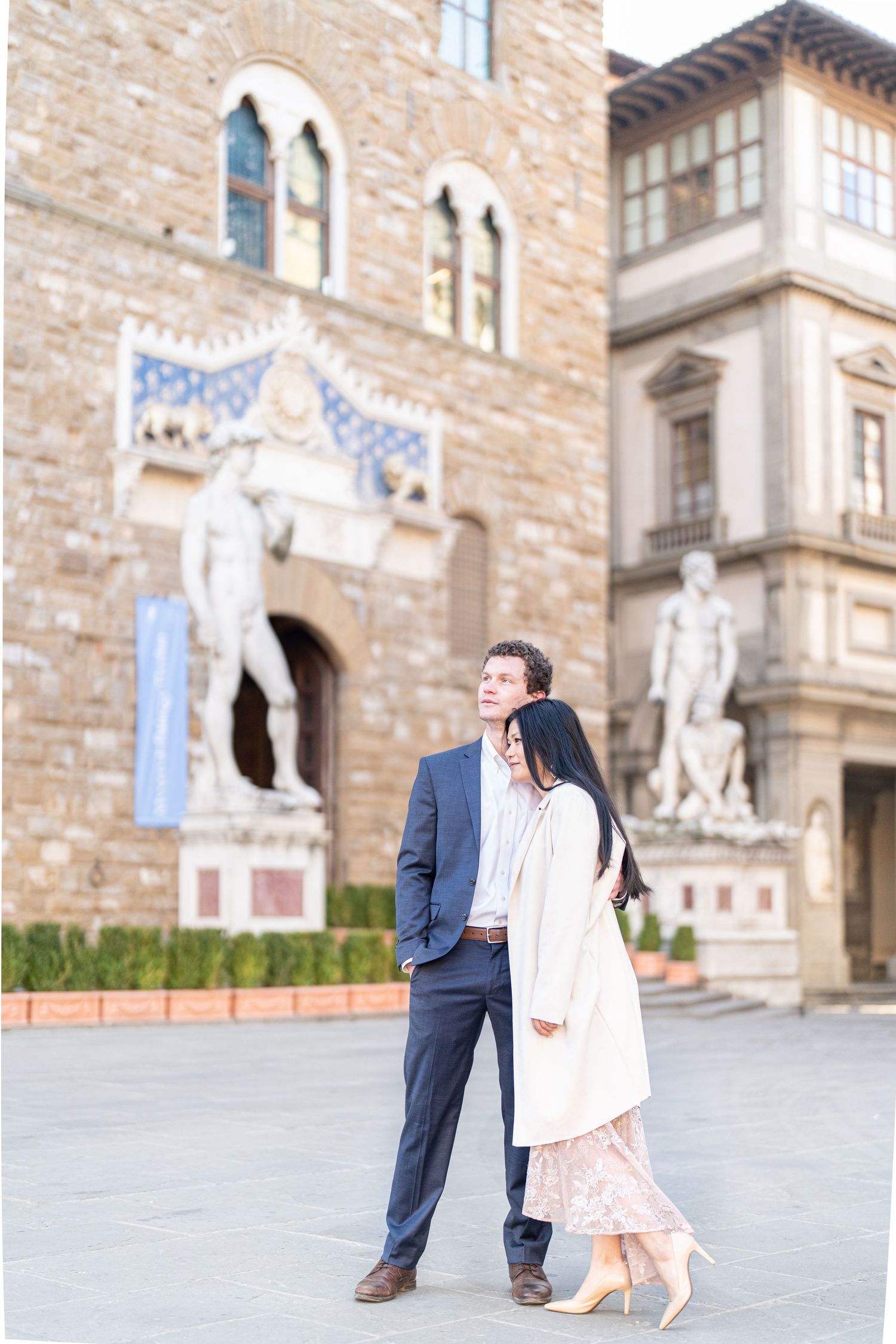 A couple in formal attire walks together outside a historic stone building with ornate architecture.