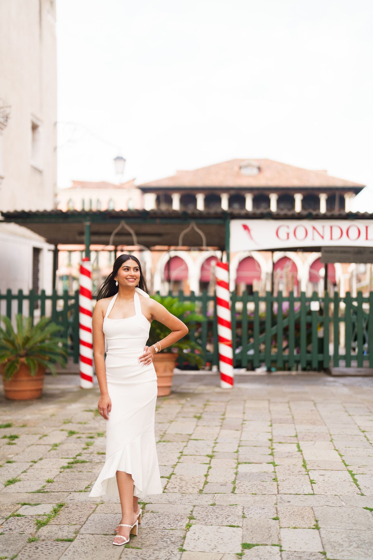 A woman in a white dress poses gracefully on a stone patio with red and white striped poles in the background.