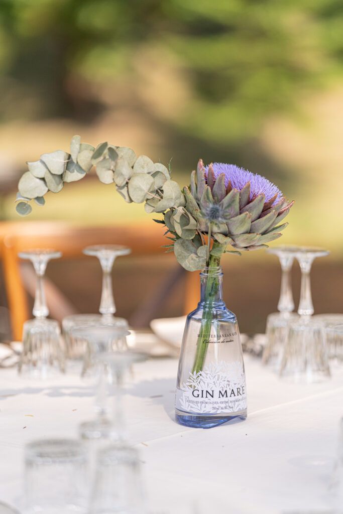 Wedding table settings with eucalyptus centerpieces and gin bottle decorations on white linens.