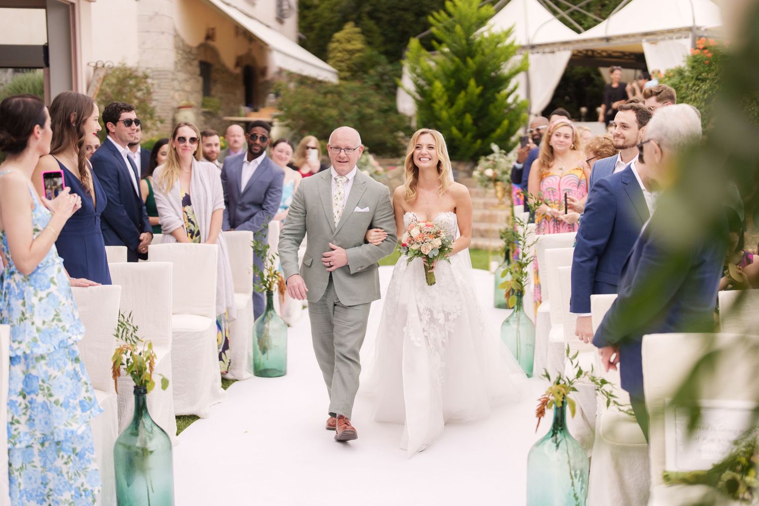 Wedding ceremony aisle decorated with blue glass bottles and white chairs as guests look on.