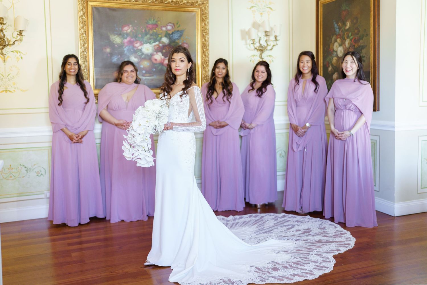 Bridal party lineup with bridesmaids in lavender dresses against white paneled walls.