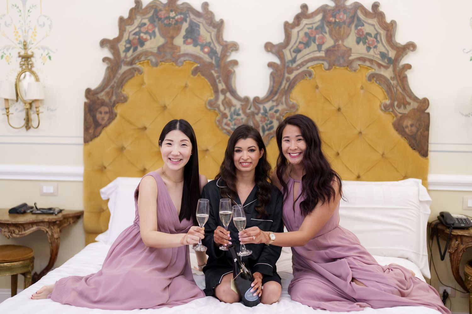 Three women in matching mauve dresses lounge elegantly on a luxurious bed in an ornate room.