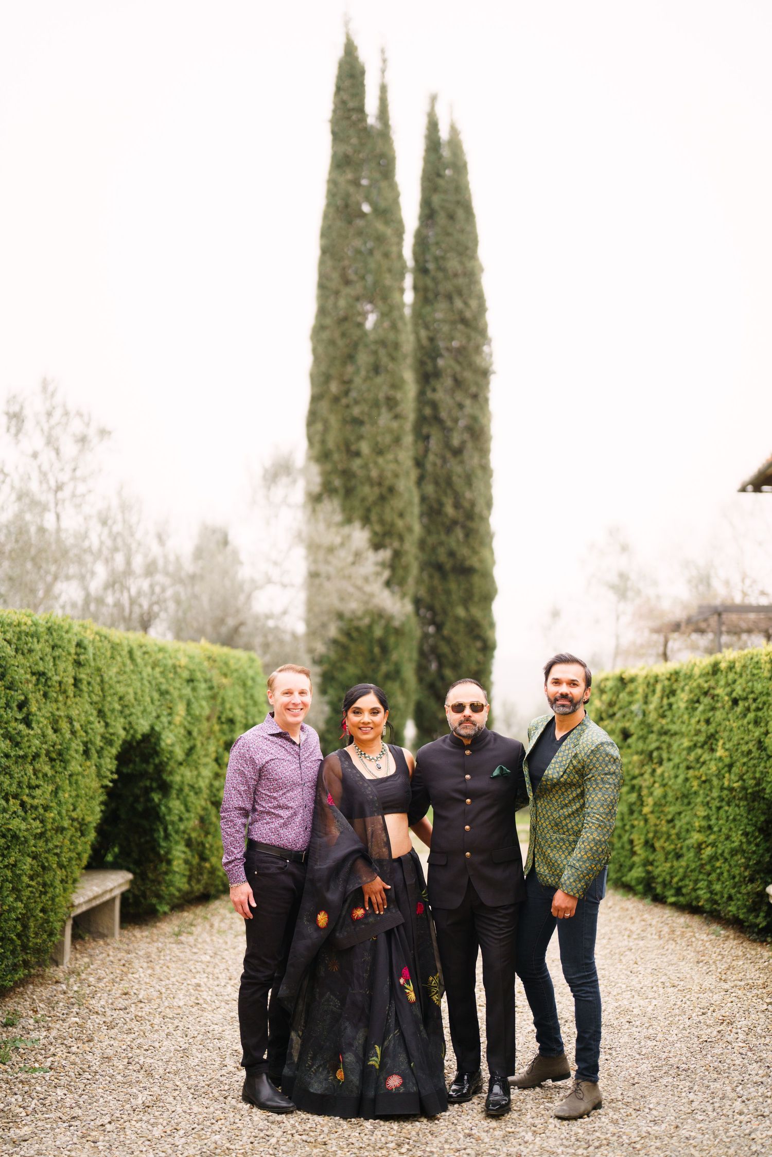 Group portrait in casual attire standing on a garden path with cypress trees and hedges.