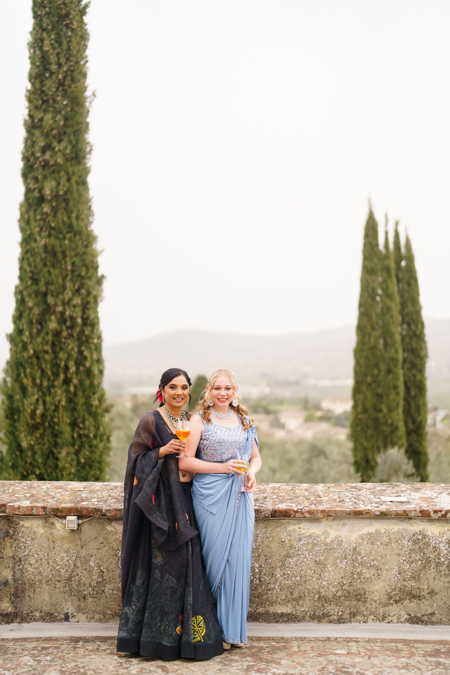 Two people in contrasting outfits pose together on a terrace with cypress trees in the background.