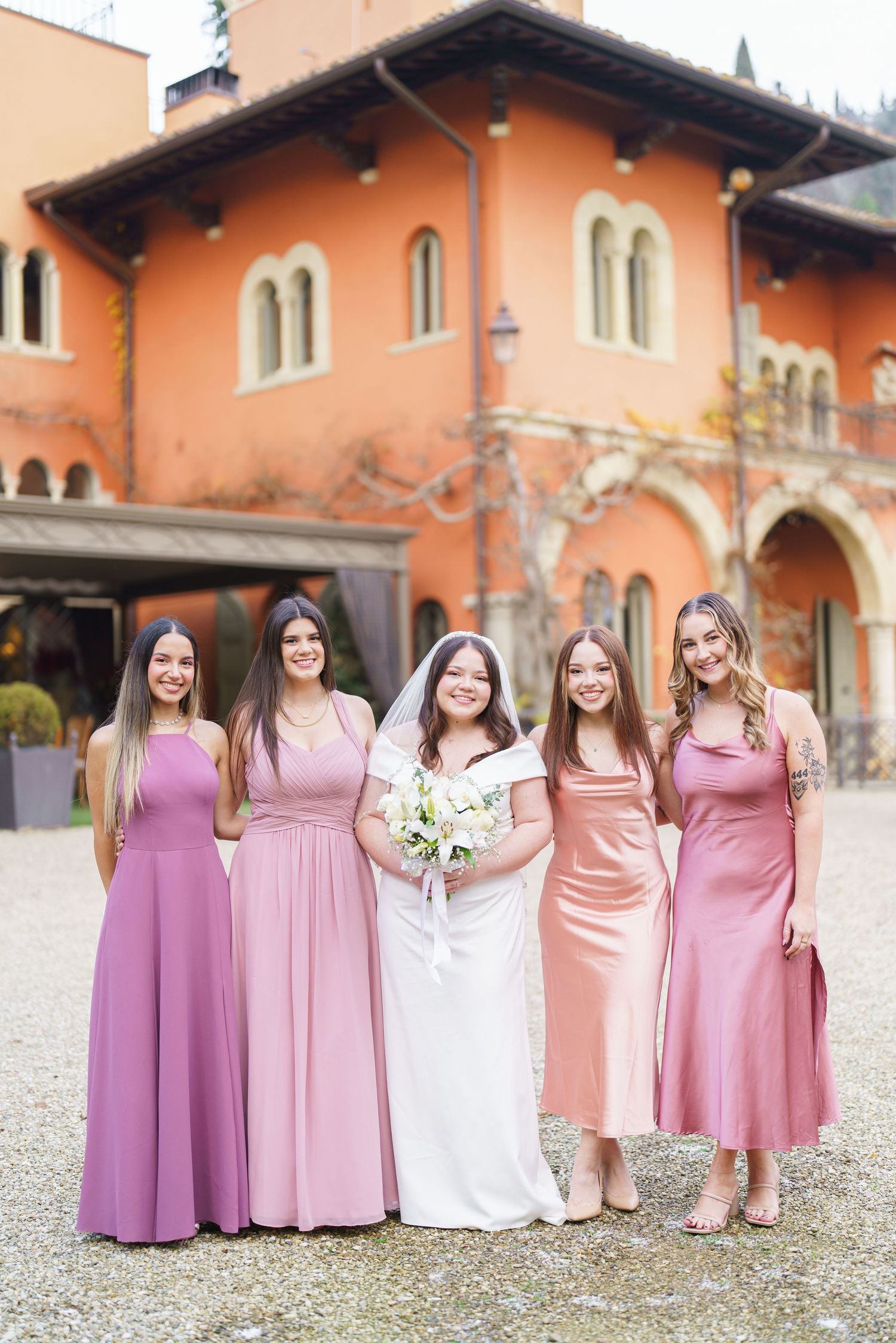 Bridal party in coordinating pink and purple dresses stands together on gravel courtyard.