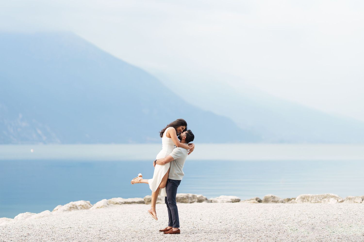A couple embraces romantically on a scenic white sandy beach with mountains and ocean in the background.