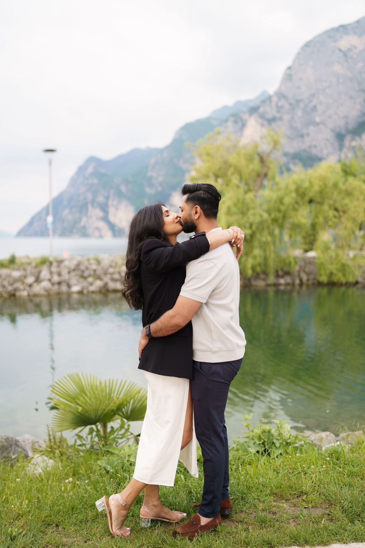 A couple shares a romantic moment by a scenic mountain lake with lush greenery in the background.