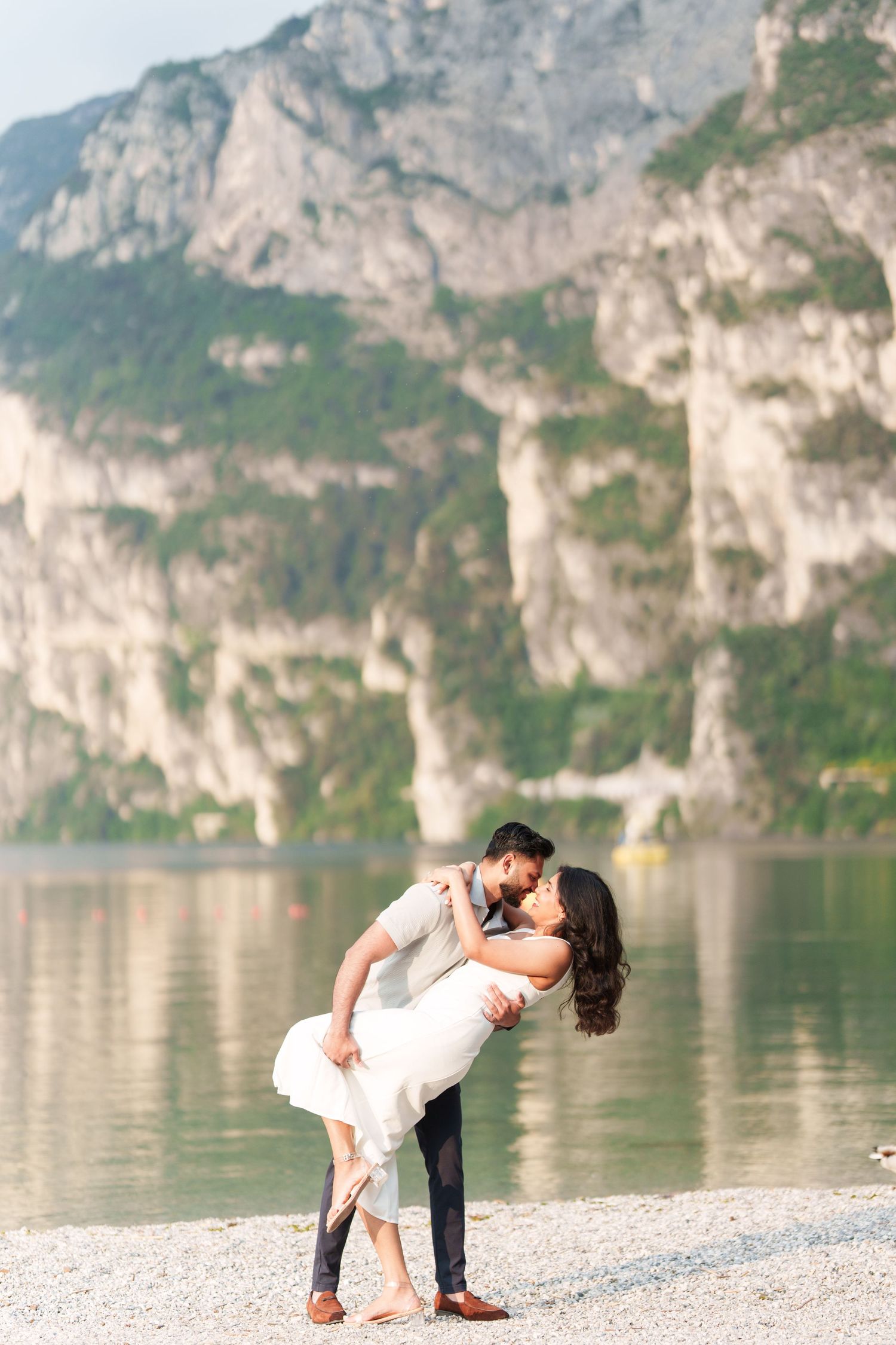 A romantic couple dips and embraces on a scenic pebble beach with towering mountain cliffs and lake in the background.