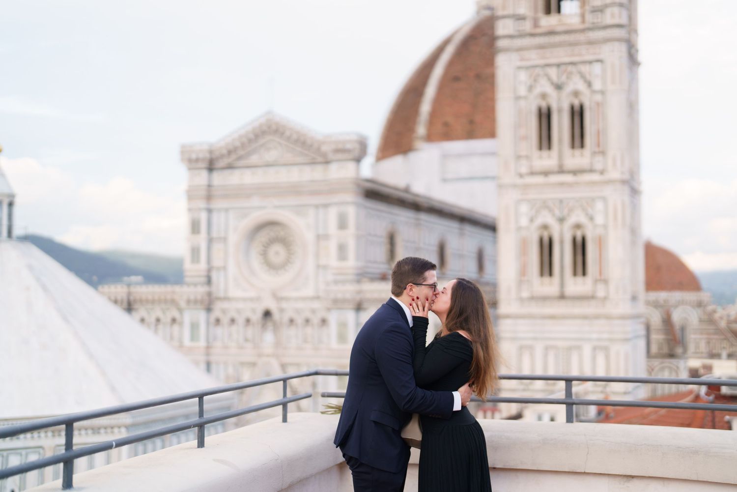 A romantic couple shares a kiss on a balcony overlooking the Florence Cathedral and historic city skyline in Italy.