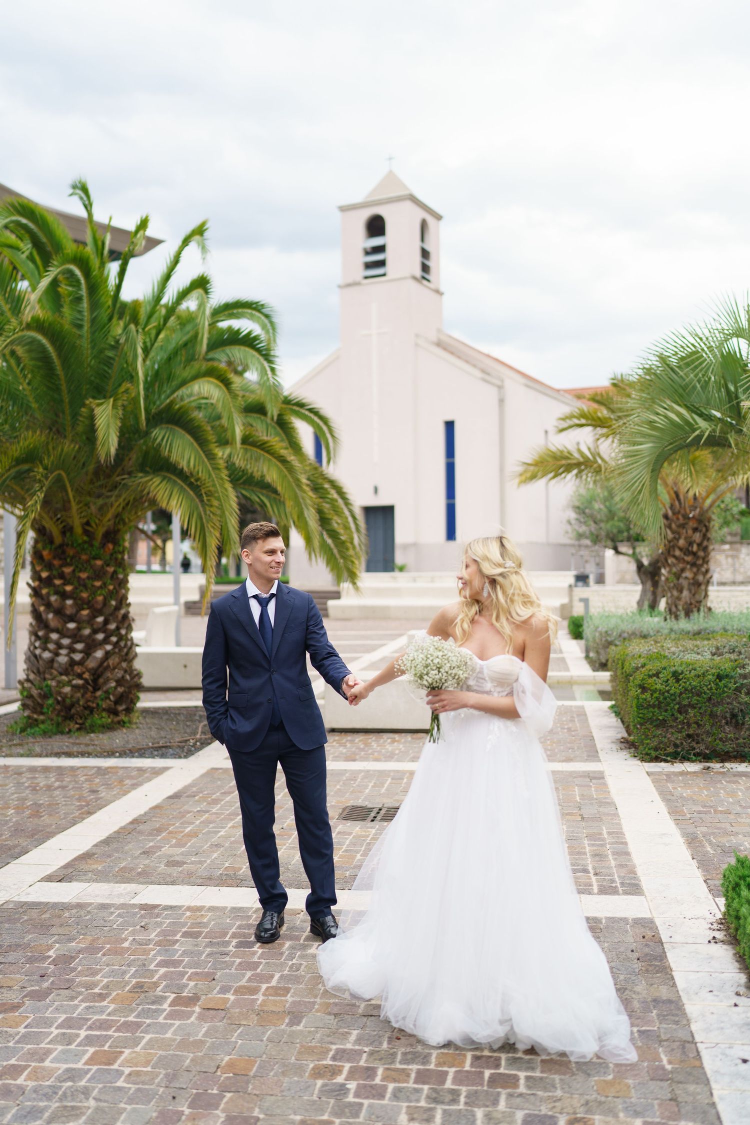 A couple in wedding attire dance and twirl together on a brick courtyard surrounded by palm trees and a white church.