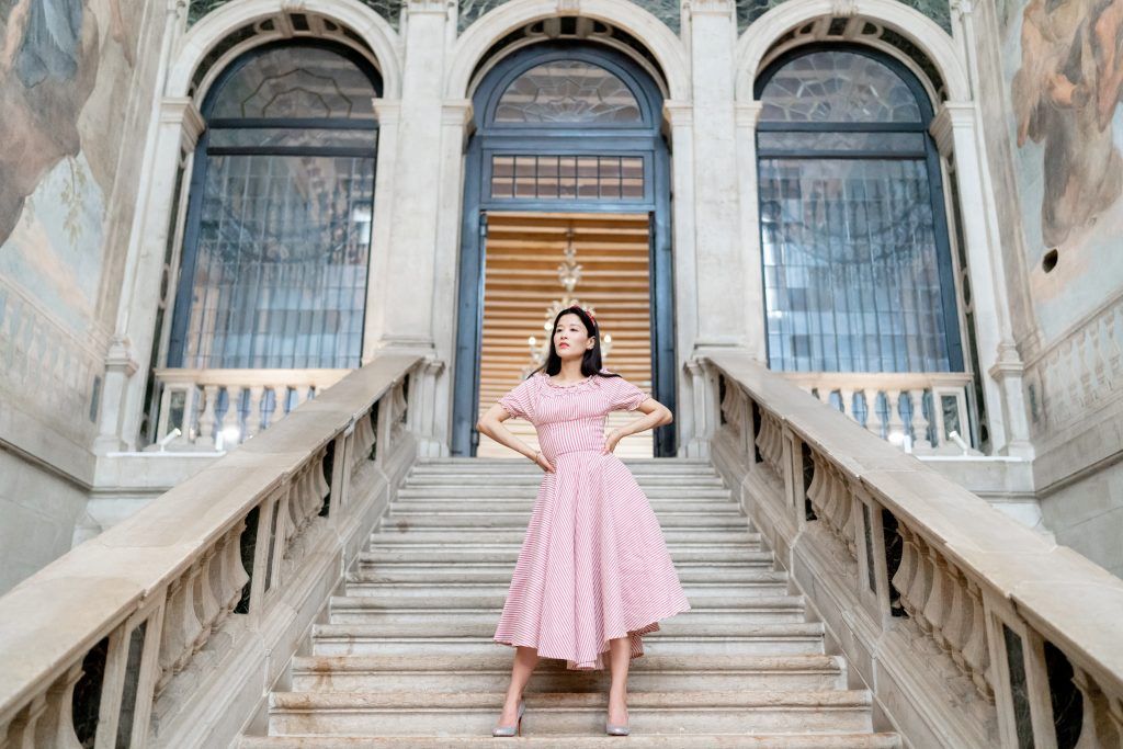 A person in a pink dress poses gracefully on a grand marble staircase in an elegant historic building interior.