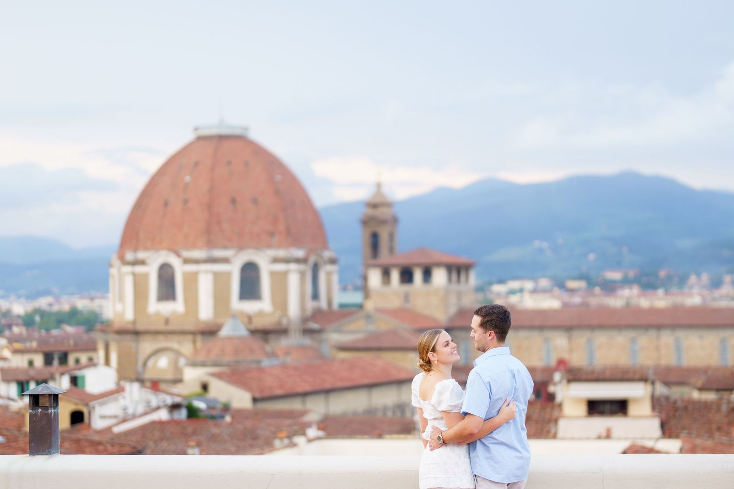 A couple shares a romantic moment on a rooftop in Florence, Italy with the iconic Duomo cathedral dome in the background.