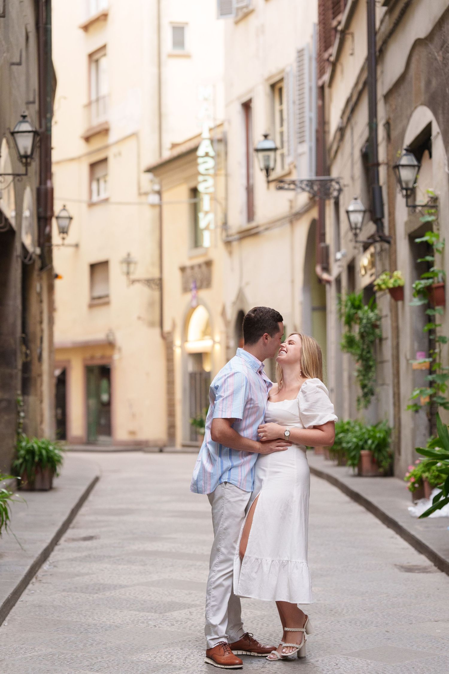 A romantic couple shares a tender moment on a charming European cobblestone street lined with historic buildings.