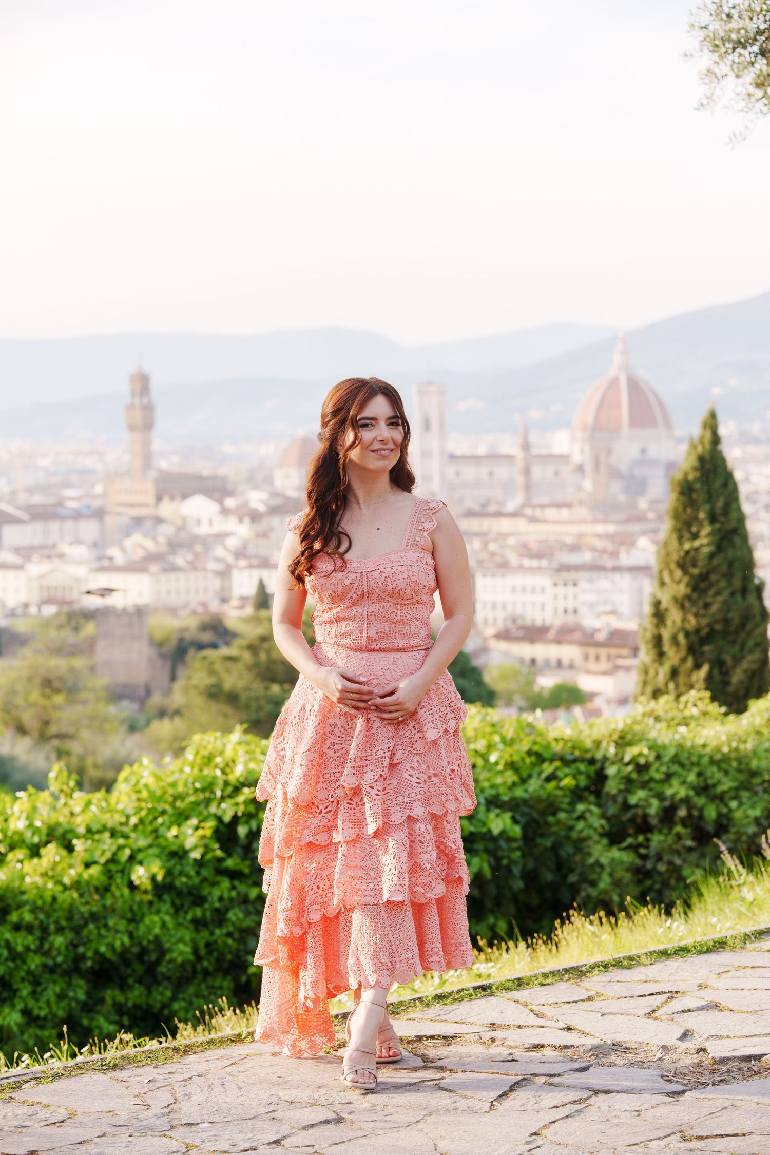 A person in a coral lace dress poses with the historic Florence skyline and Duomo cathedral visible in the background.