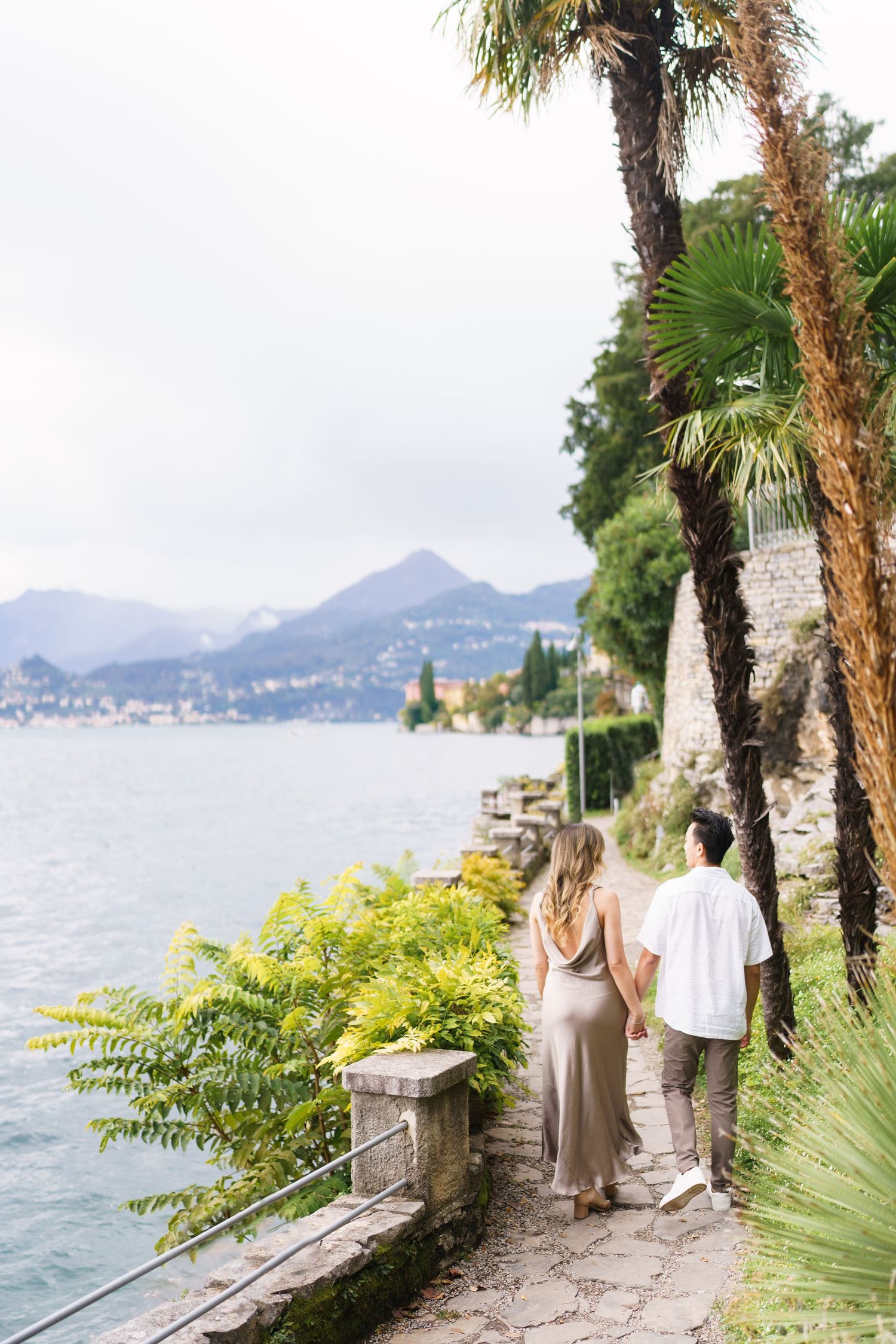 A couple strolls along a scenic lakeside path with palm trees and mountains in Lake Como, Italy.