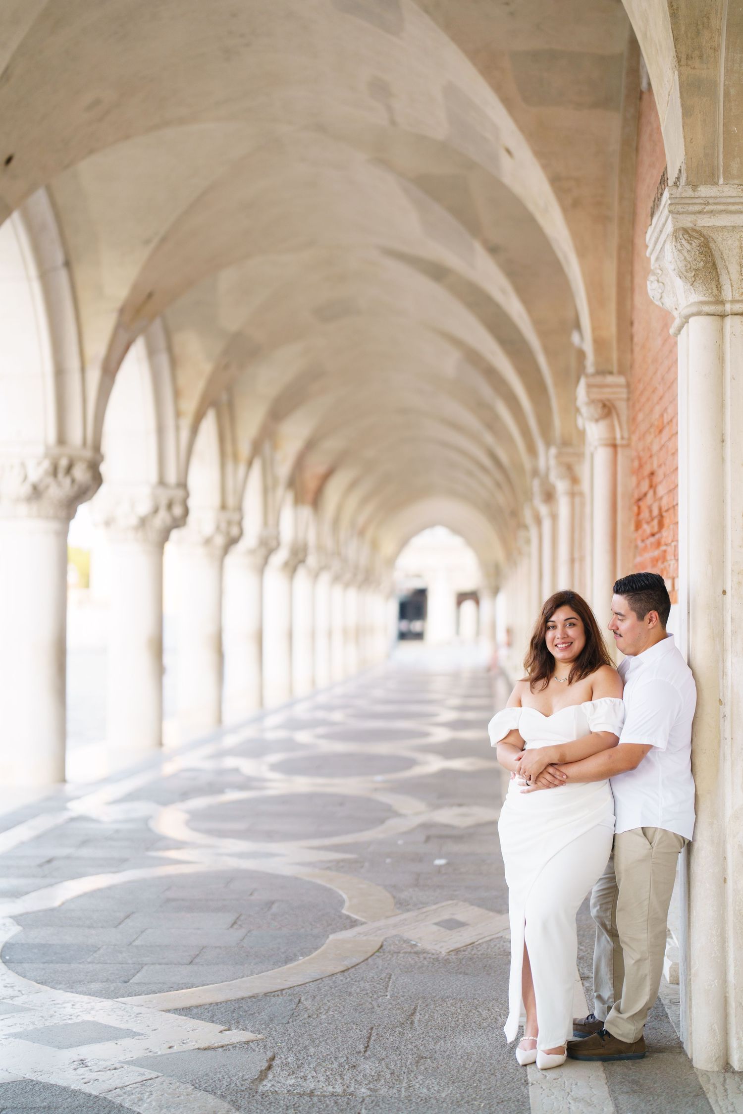 A couple in white outfits poses under an elegant arched colonnade with repeating architectural columns.