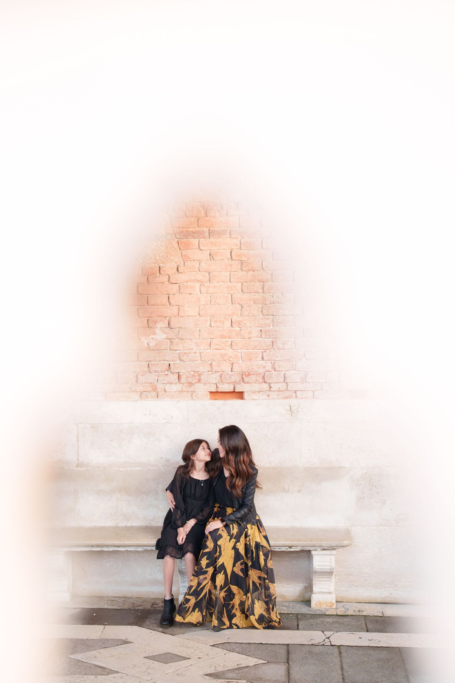 Two people in matching floral dresses sit together on white stone steps against a brick wall.