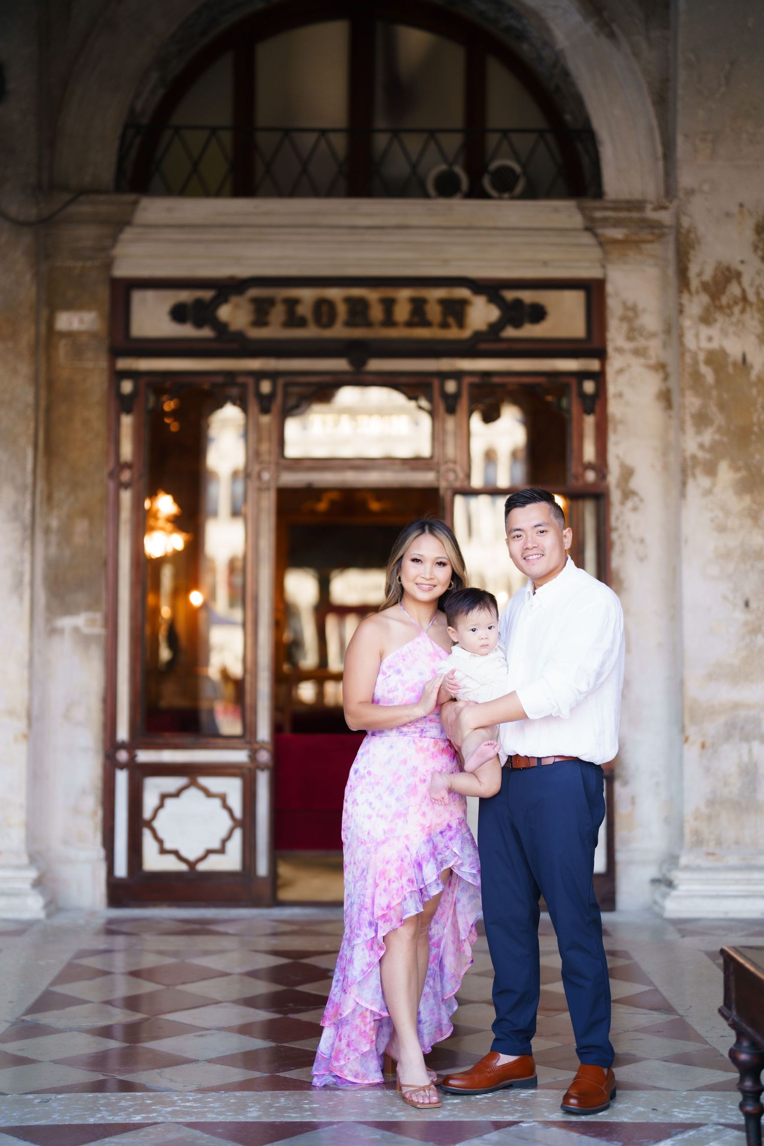 A young Asian family poses in front of Caffe Florian in Venice, Italy, parents holding their baby.