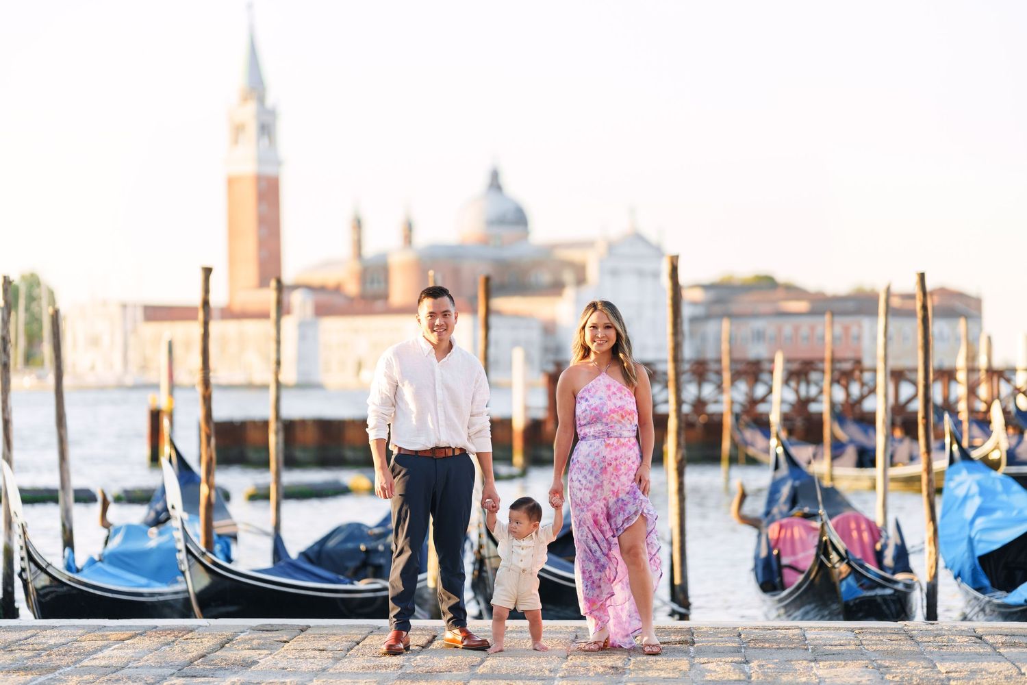 Family of three posing on Venice waterfront with gondolas and San Giorgio Maggiore church in golden hour light.